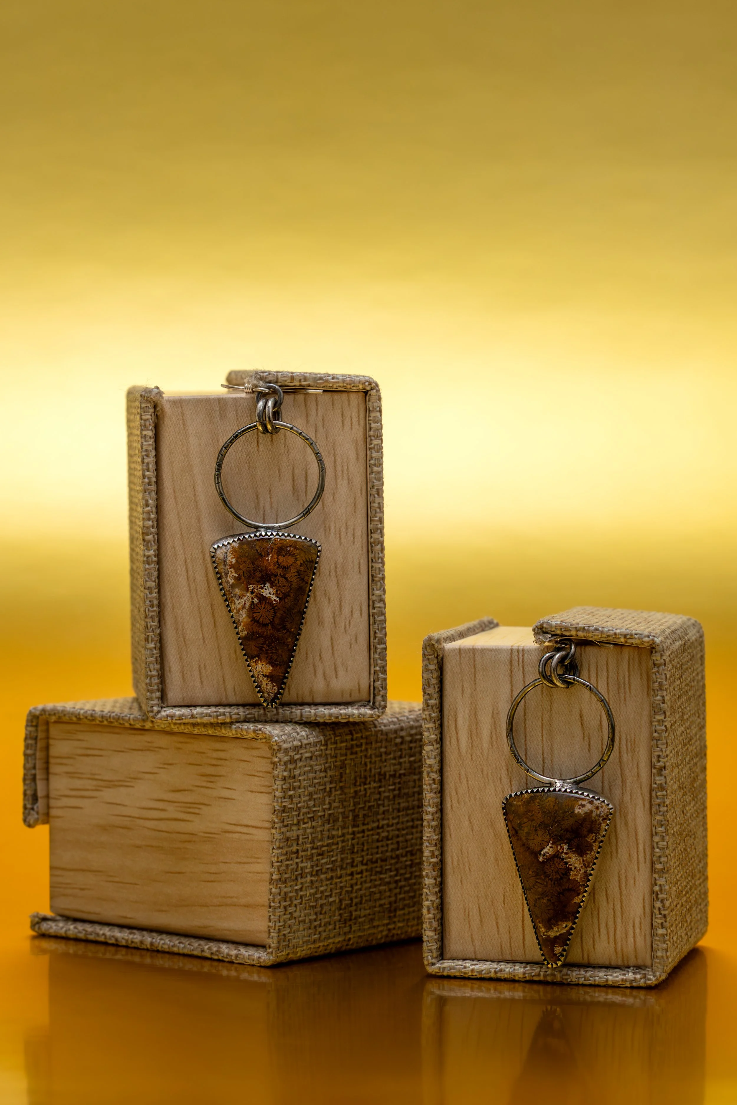 Two jewelry pieces featuring amber stones, each hanging from a ring, displayed on wooden and woven boxes against a yellow background.