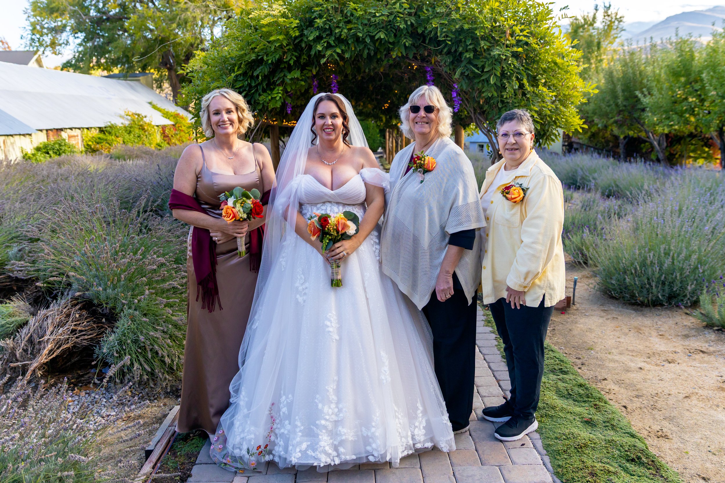 Four women standing on a brick pathway in a garden with purple flowers and green trees; a bride wearing a white wedding gown and veil holds a bouquet, flanked by women in casual and semi-formal attire, all holding flower bouquets.