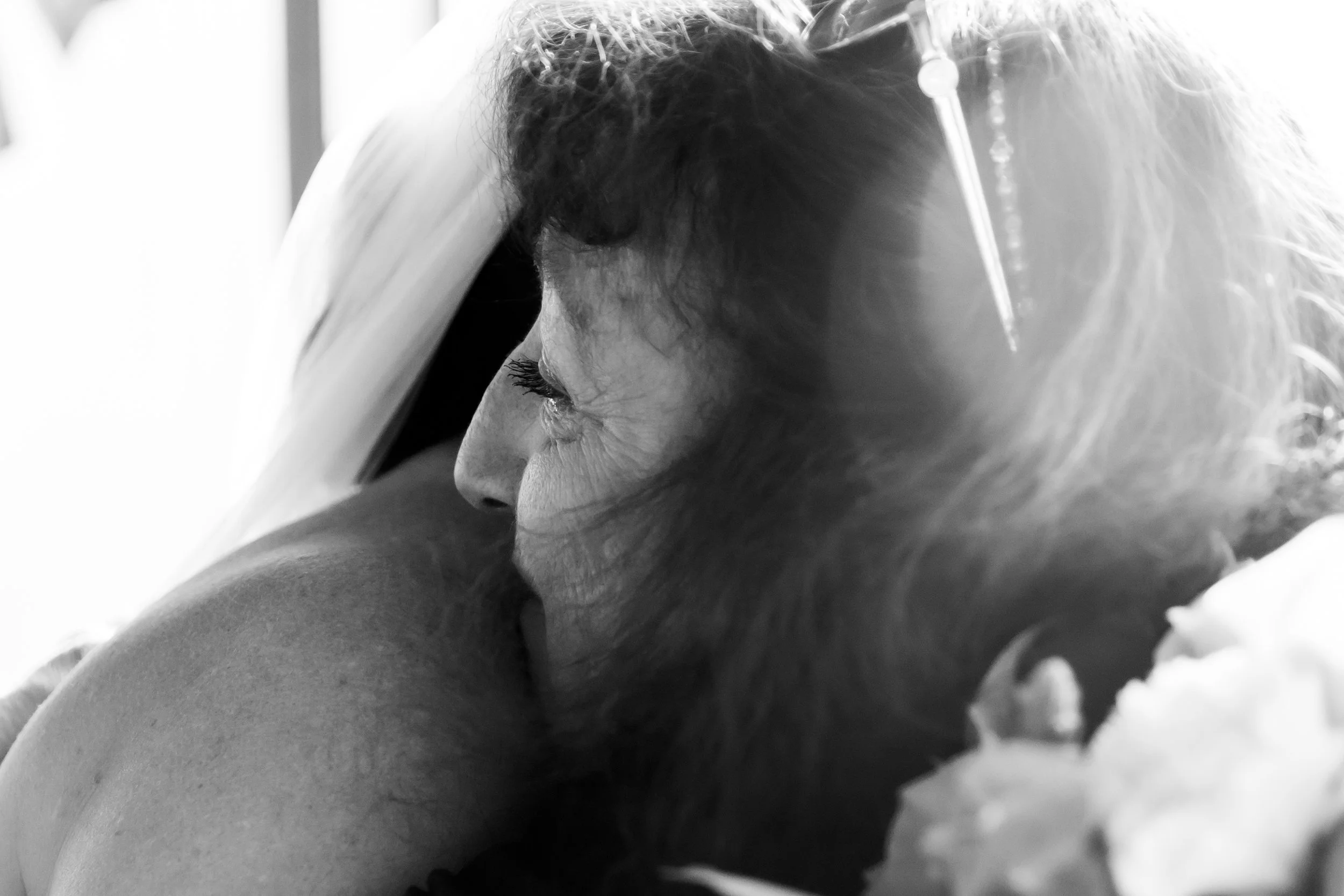A woman with curly hair and long eyelashes lies on her side, resting her head and arm on a pillow, with a medical IV needle in her hand, in a grayscale photo.