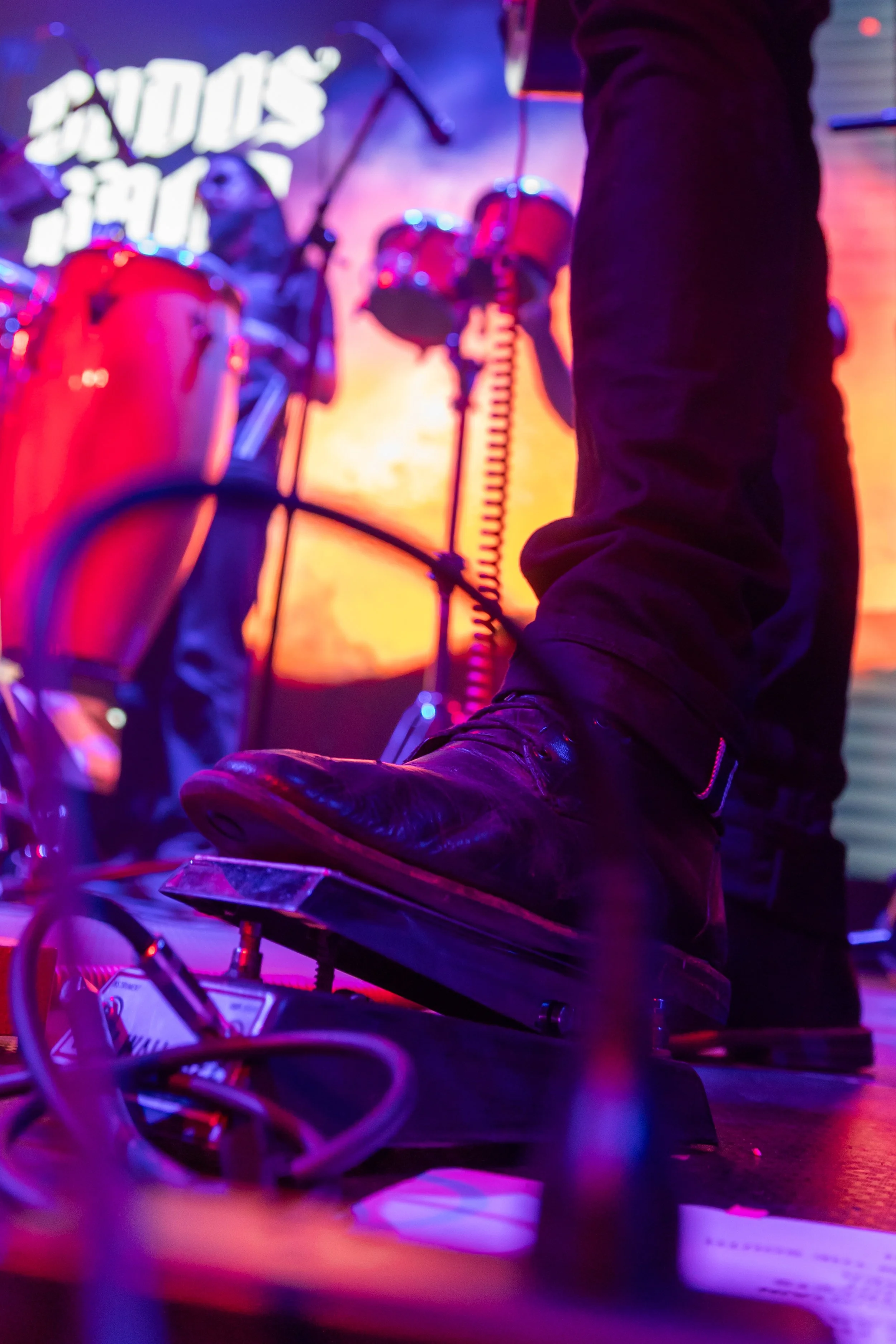 Close-up of a musician's foot on a pedal, with other band members playing drums in the background on stage during a live performance.