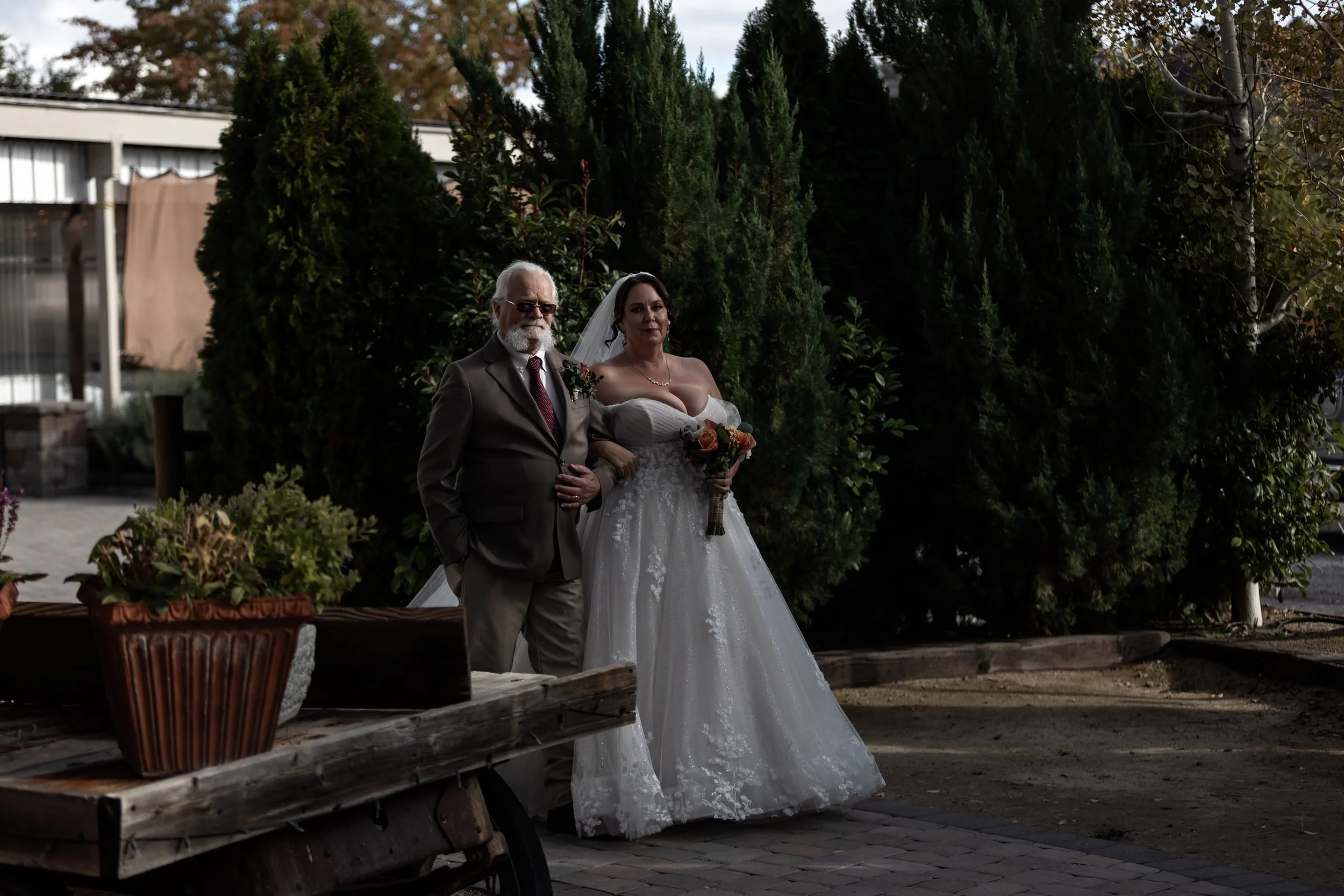 A bride in a white wedding dress holding a bouquet and an older man in a suit with sunglasses standing outdoors near tall greenery with trees and a building in the background.