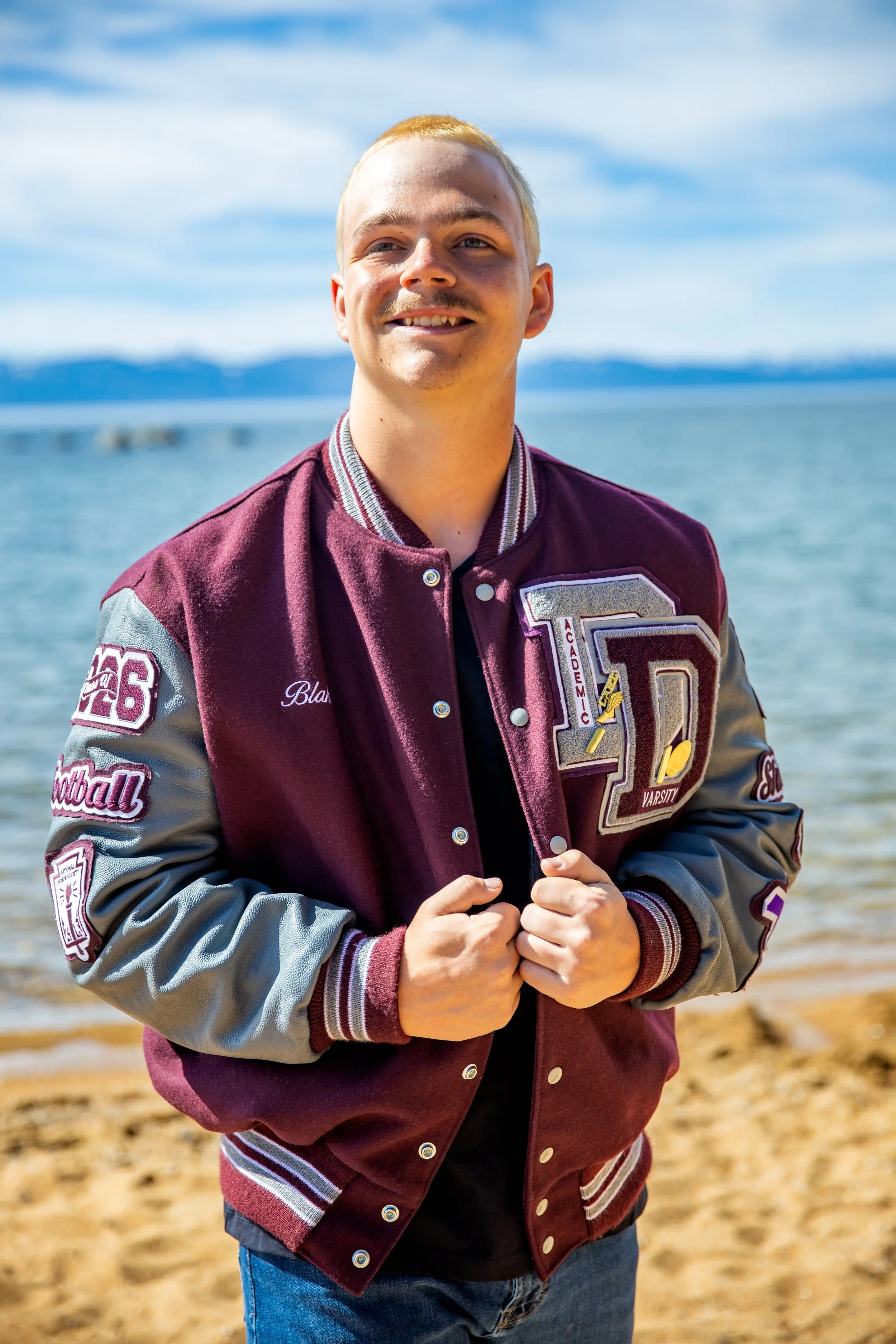 Young man on beach, wearing a maroon and grey varsity jacket, smiling, with water and sky in background.