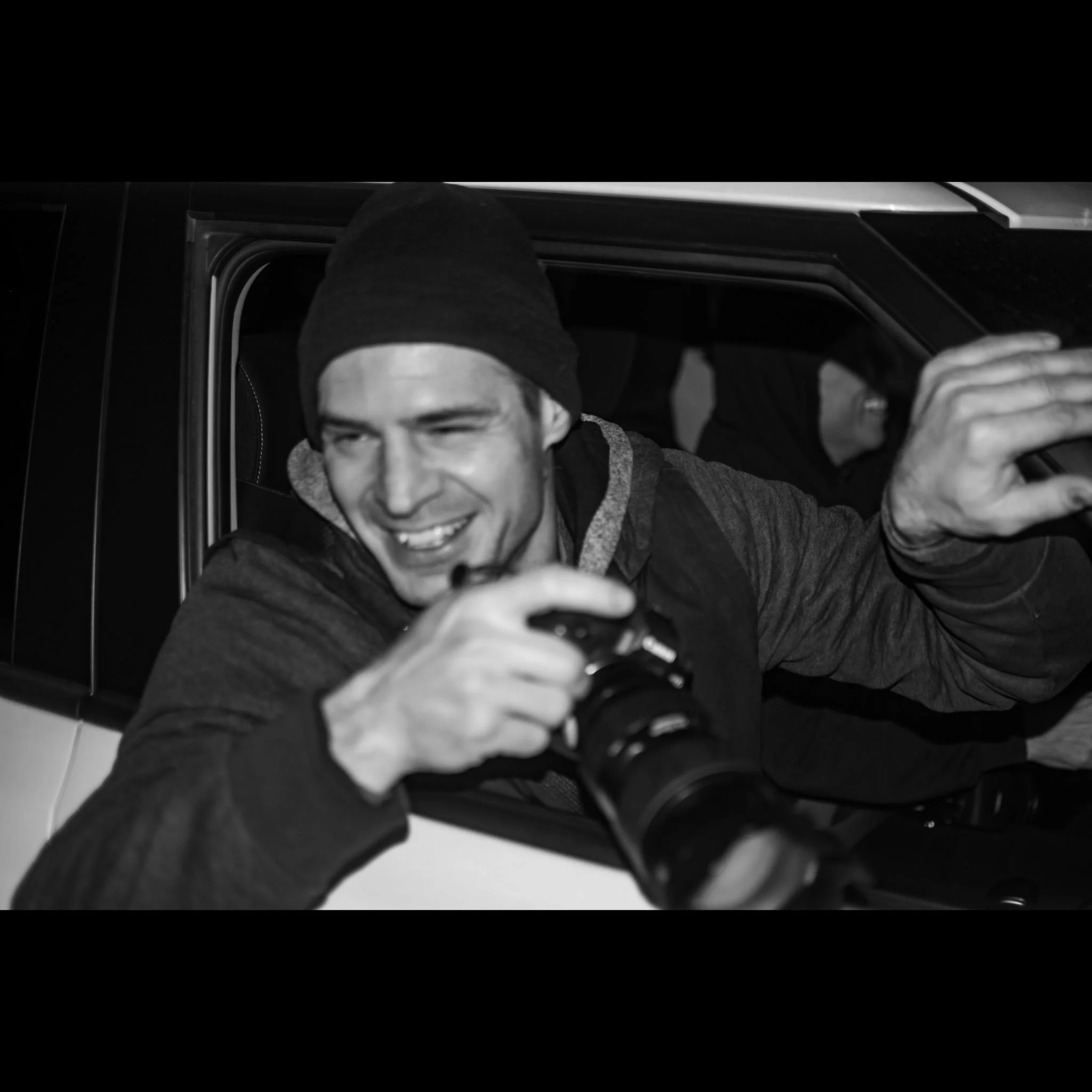A young man wearing a hoodie and black beanie taking a selfie inside a car, with his face partially visible and a smile, reflected in a smartphone.