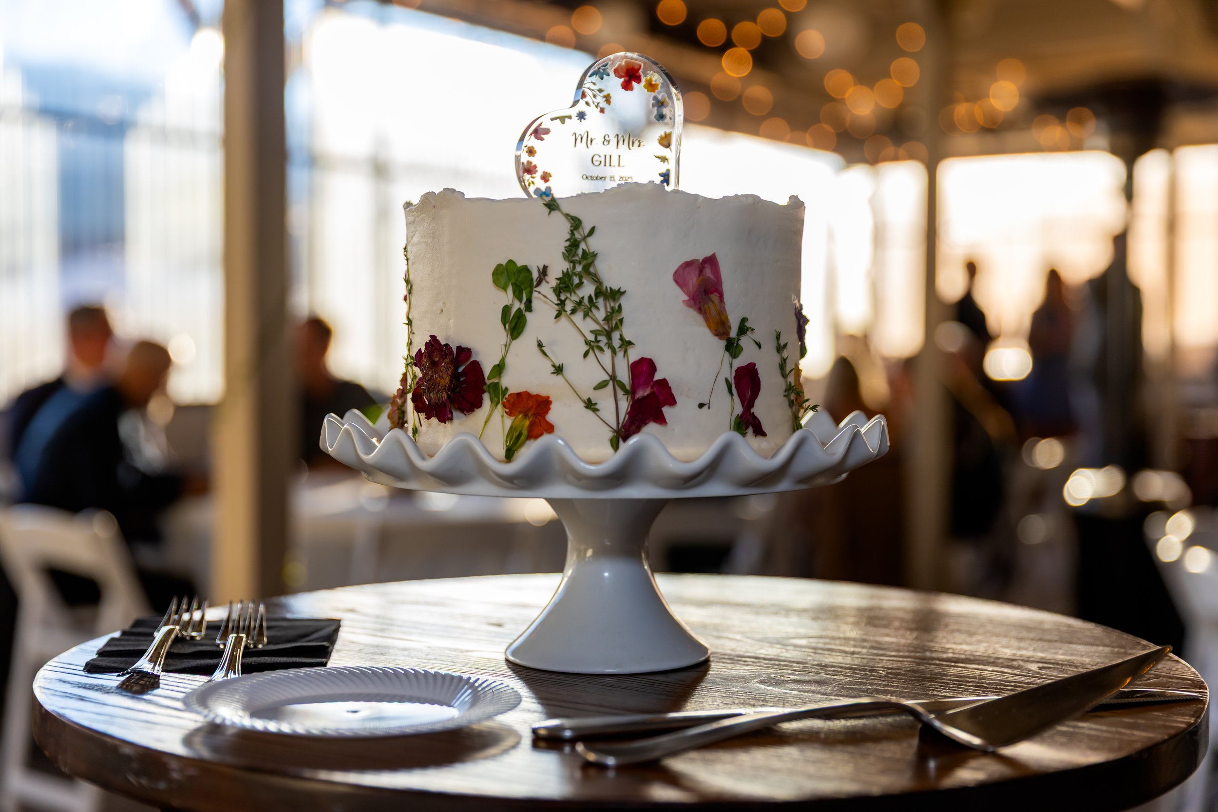 A square wedding cake decorated with edible flowers and greenery, topped with a heart-shaped topper that reads 'Mr. & Mrs. GILL' and the date October 12, 2023, placed on a white cake stand on a wooden table.