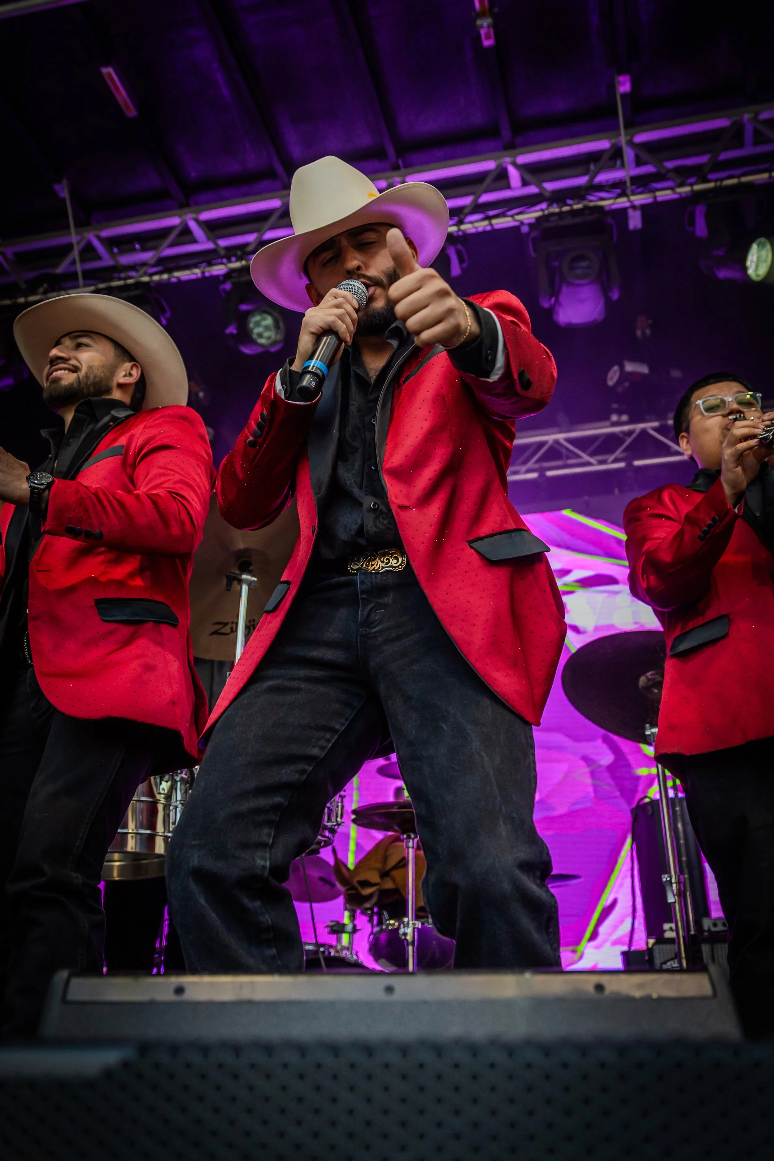 A male singer wearing a white cowboy hat, red blazer, black shirt, and black jeans, sings into a microphone while giving a thumbs-up gesture. Two other men in similar red blazers and cowboy hats are visible in the background, one playing a trumpet. T
