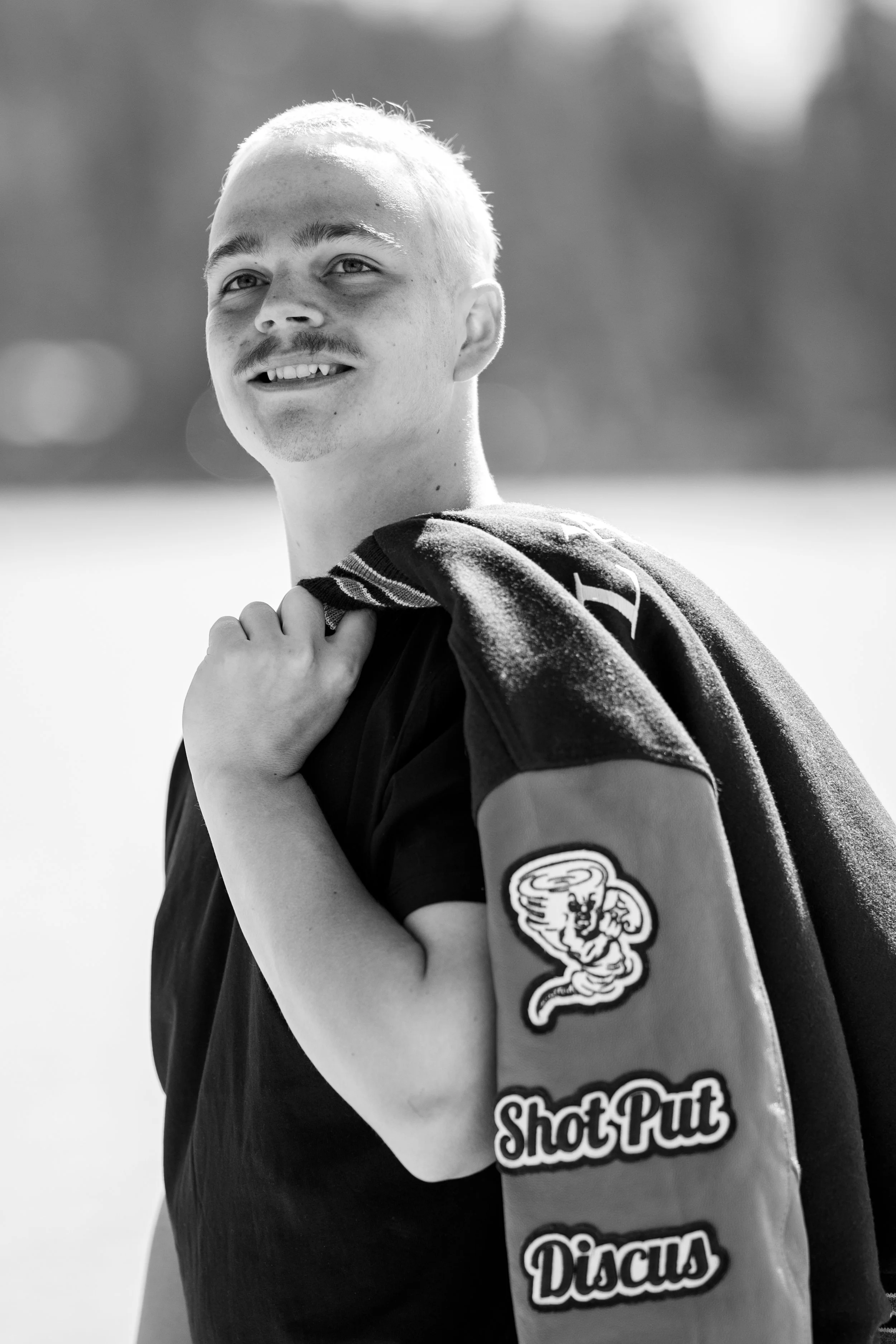 Black and white photo of a young man with short light hair, a mustache, and a gap between his front teeth smiling and looking to the side. He is wearing a sports jersey with patches and logos, and is holding a towel over his shoulder.