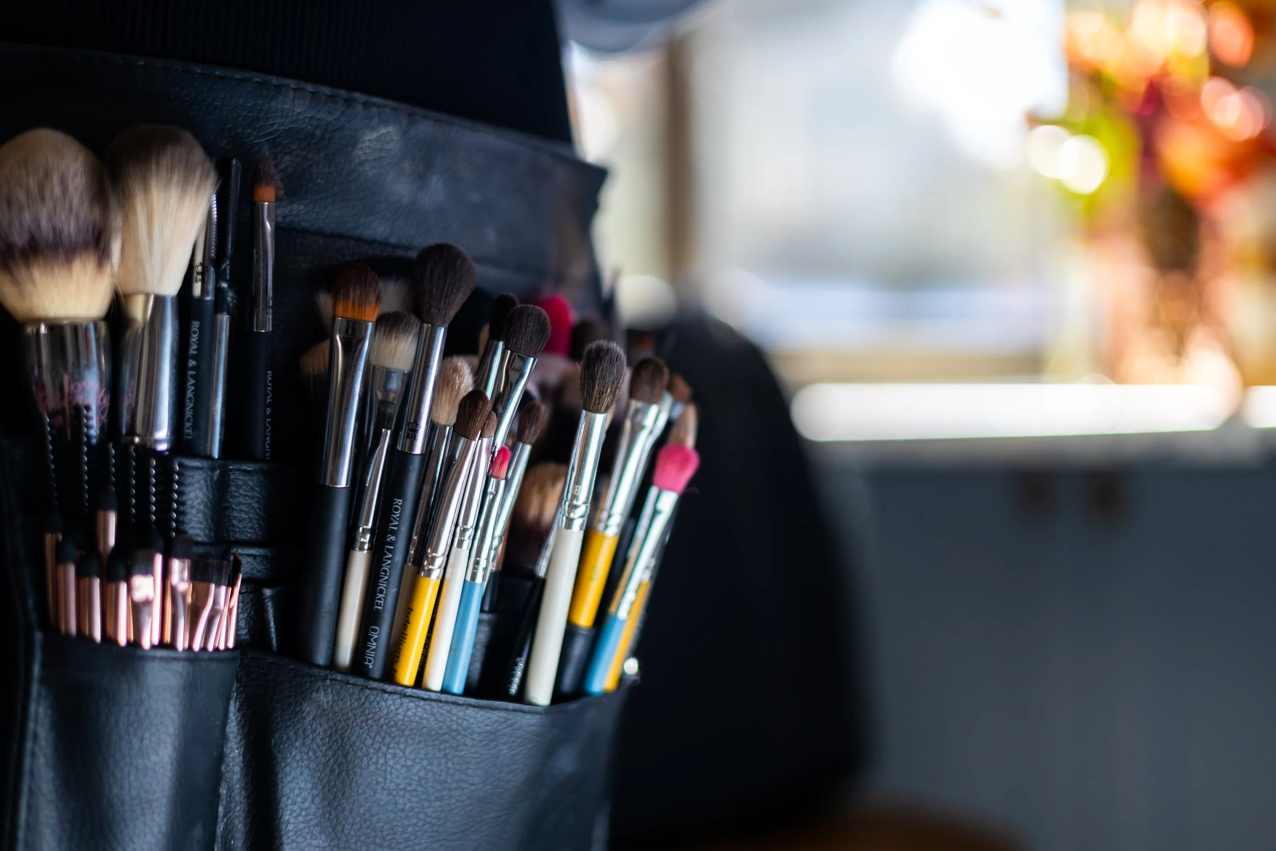 A black leather pouch with various makeup brushes inside, some with pink, brown, or black bristles, placed near a window with blurred colorful background.