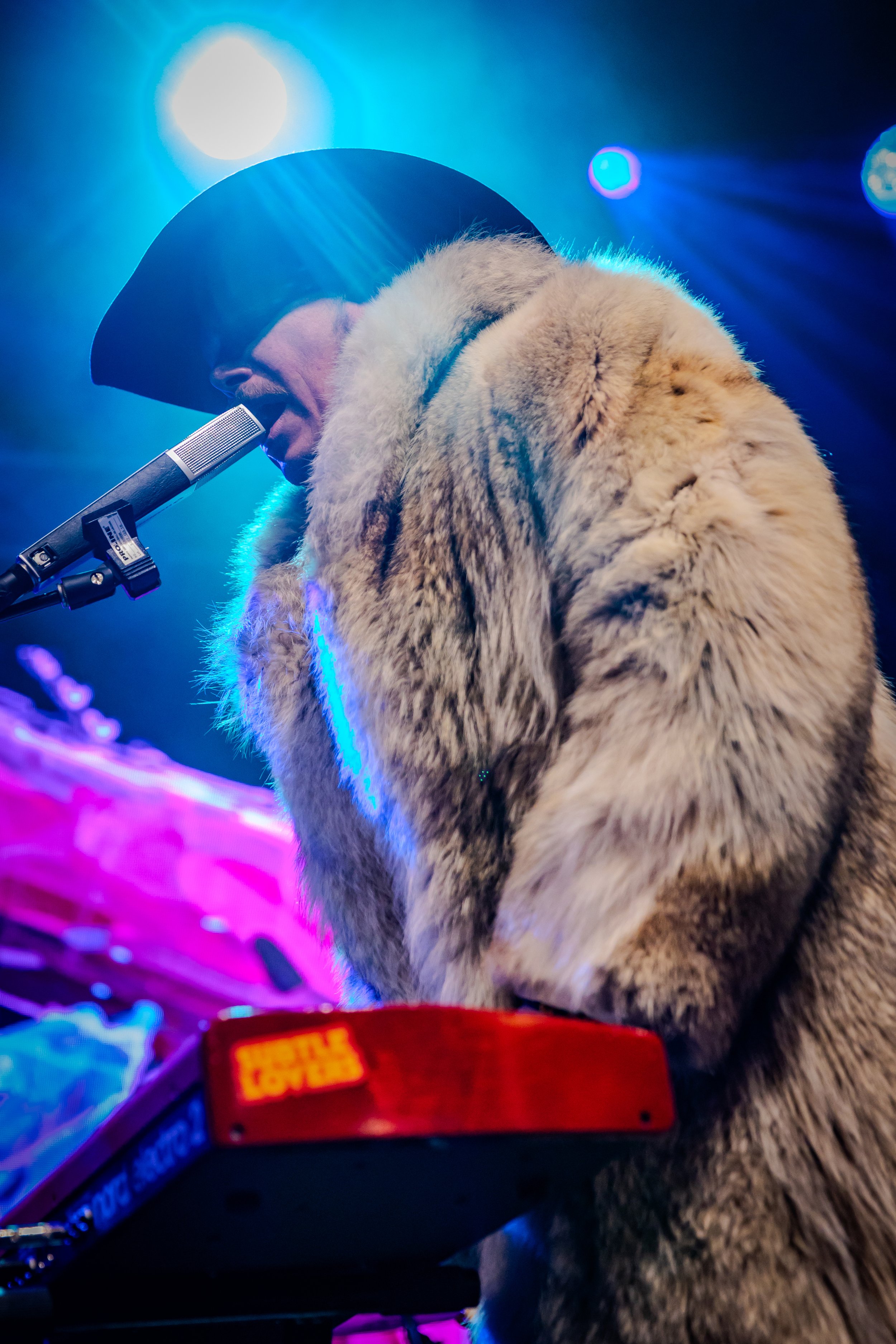 Close-up side view of a person wearing a black hat and a furry coat, holding a microphone, with colorful stage lights in the background.