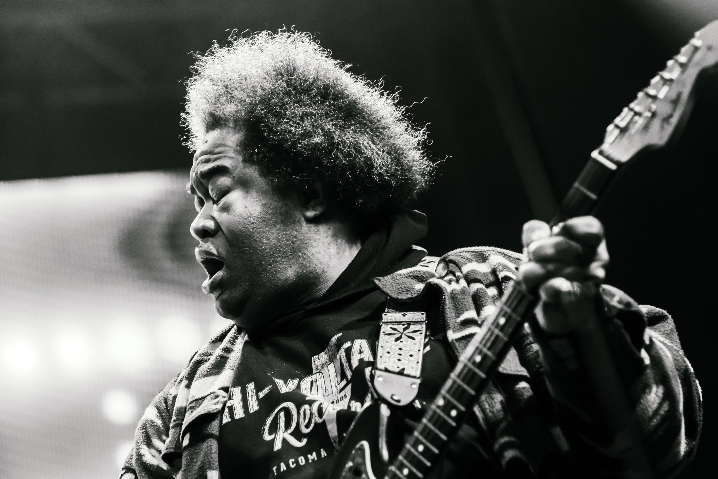 A black and white photo of a person with curly hair playing an electric guitar, eyes closed, wearing a patterned jacket.