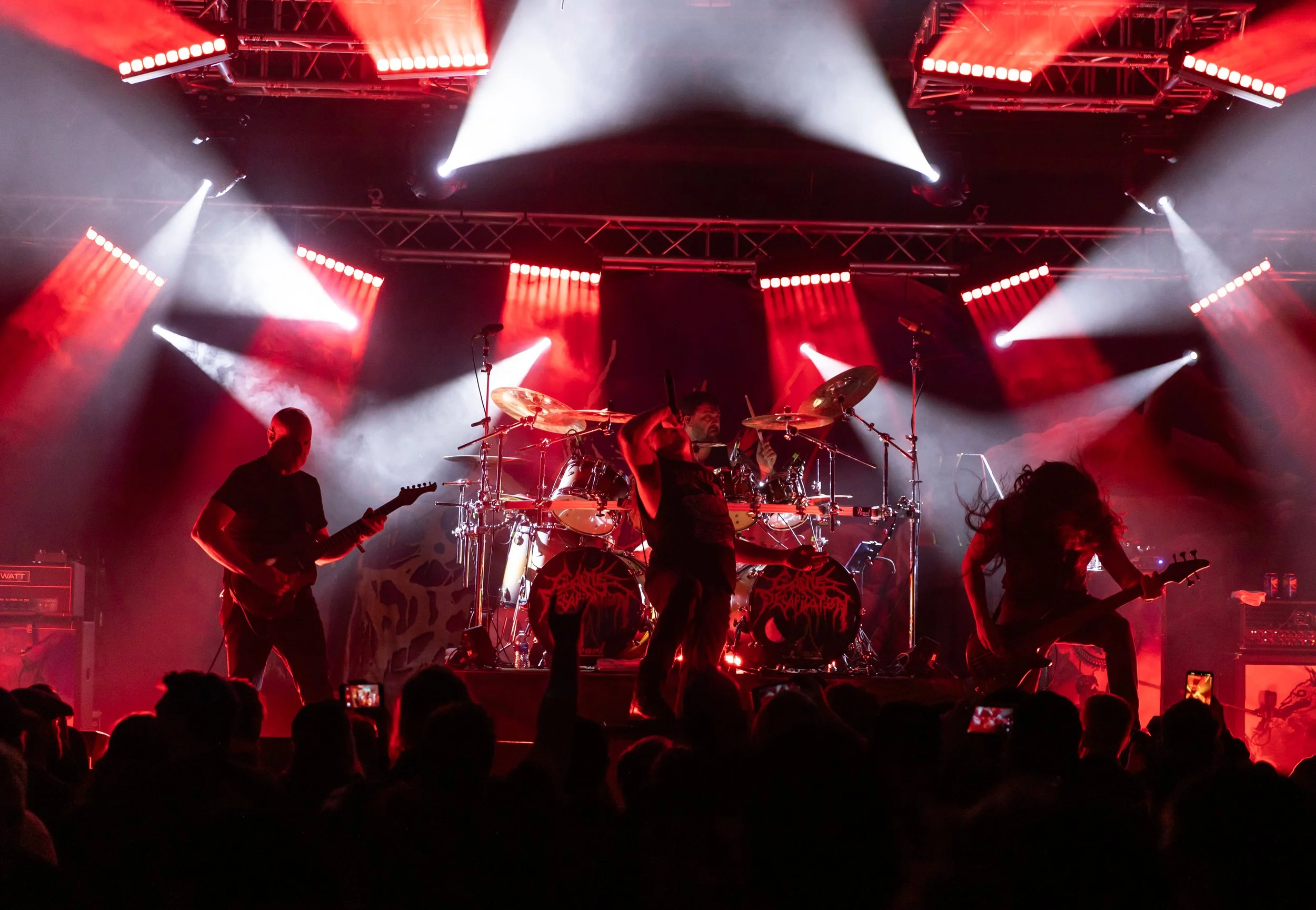 A live concert scene with a band performing on stage under bright red and white lights, with people in the audience taking photos.