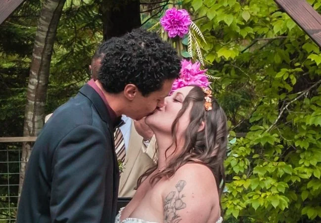 A couple kissing during a wedding ceremony with a flower arch in the background.