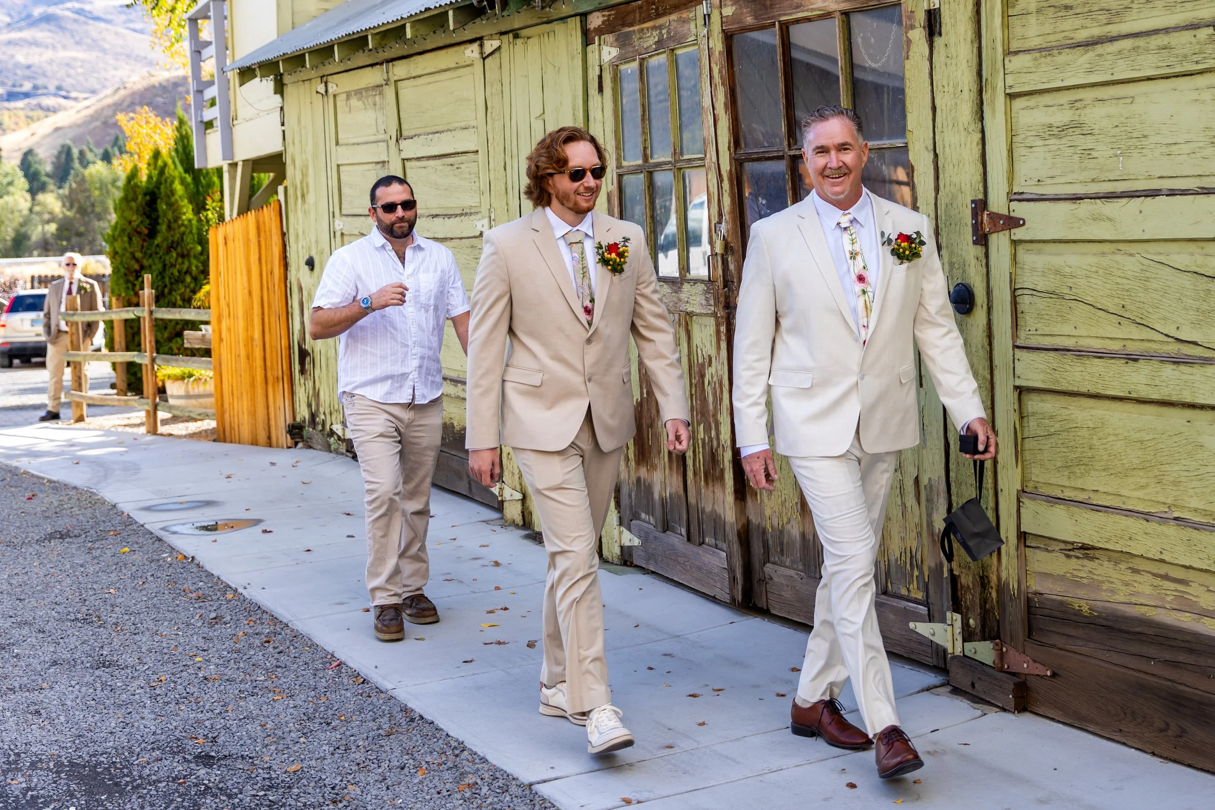 Four men walking along a sidewalk next to a weathered green building, with two wearing cream-colored suits and sunglasses, one in a casual white shirt, and the fourth in a tan suit in the background. The man in front is smiling and holding a black ba
