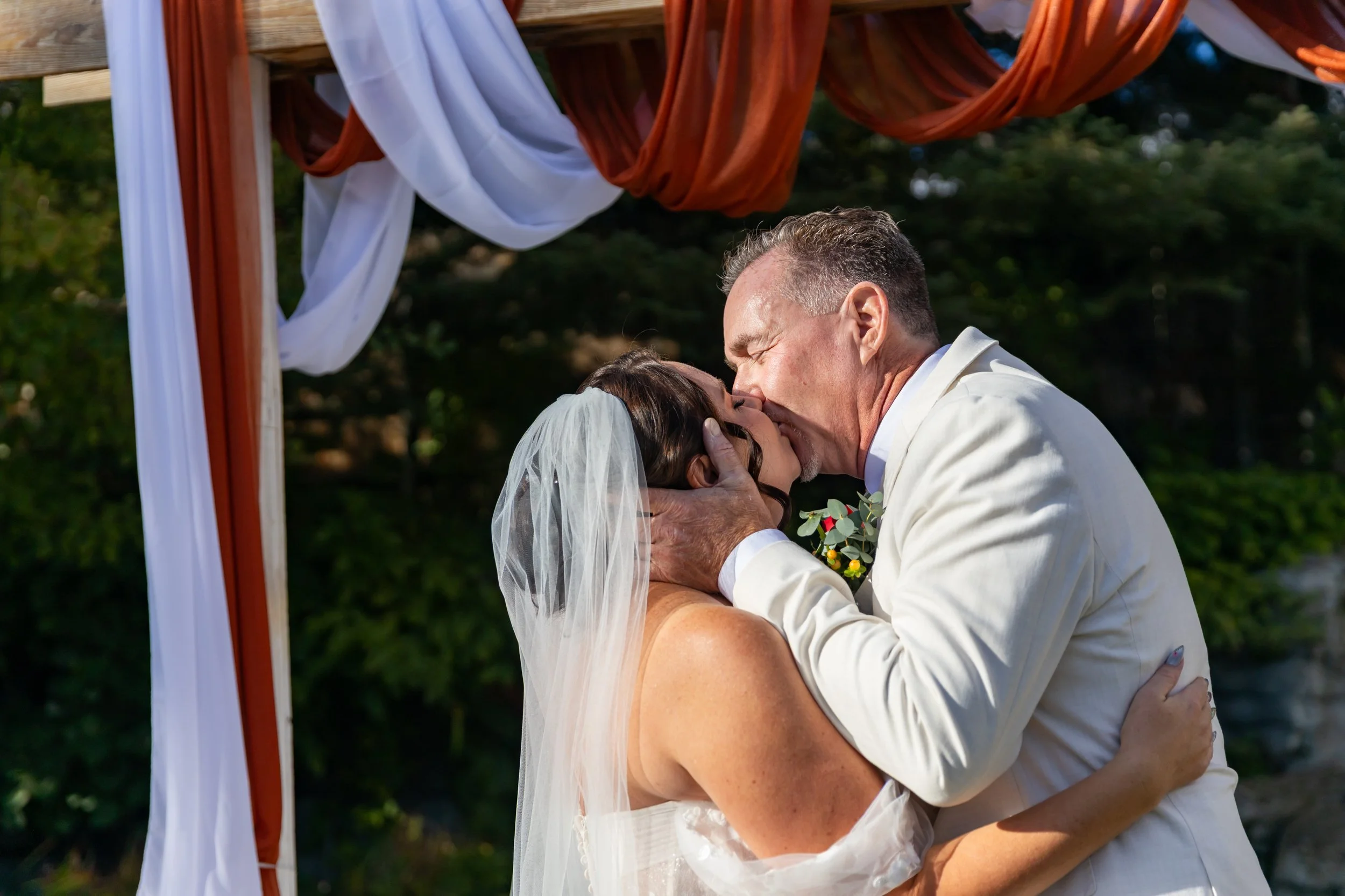 A bride and groom share a kiss during their outdoor wedding ceremony, with a decorated wooden arch draped in white and rust-colored fabric behind them.