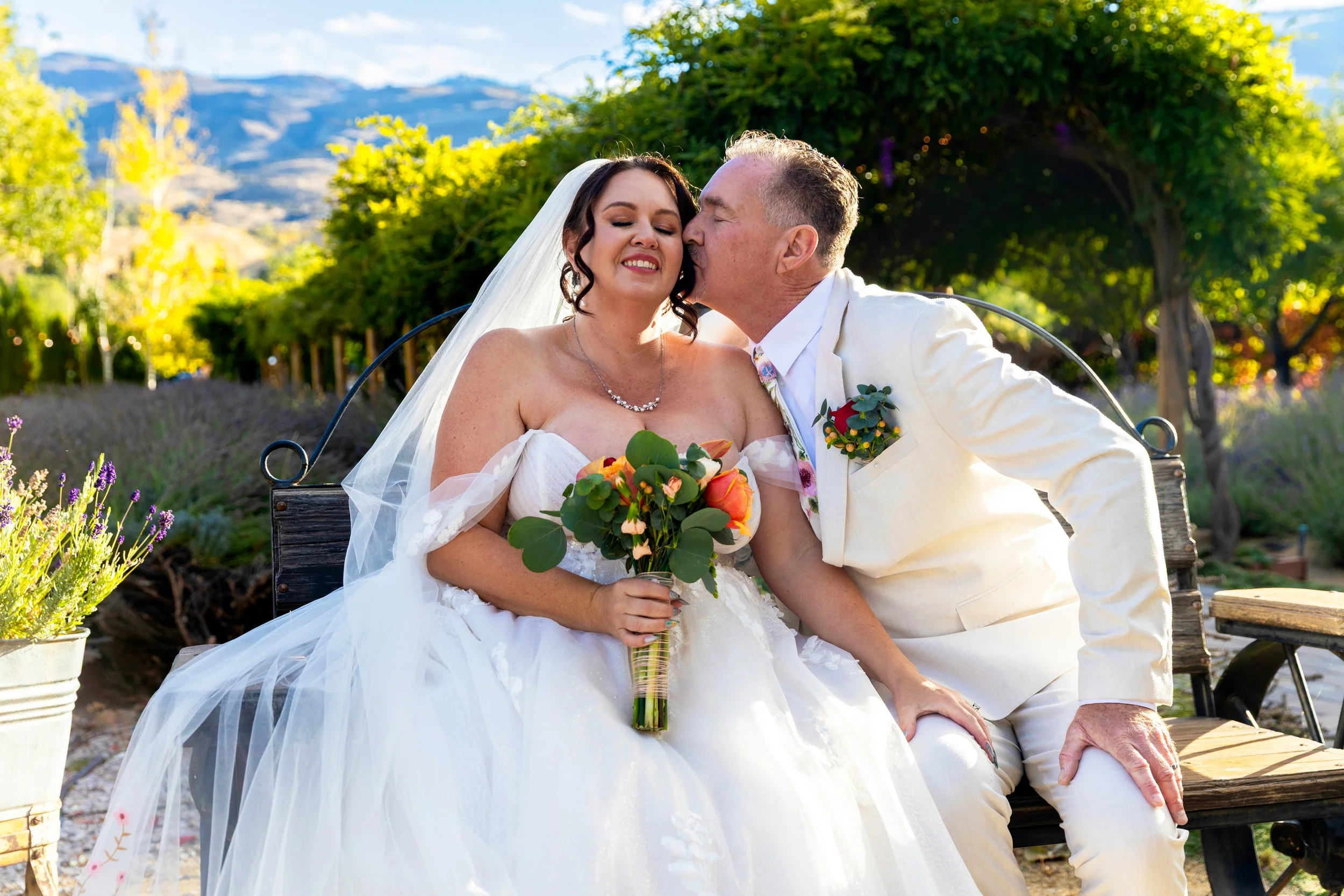 A bride and groom sitting on a wooden bench outdoors, the groom kissing the bride on the cheek. The bride is holding a bouquet, wearing a white wedding dress and veil; the groom is in a white suit with boutonniere. Green trees, colorful flowers, and 