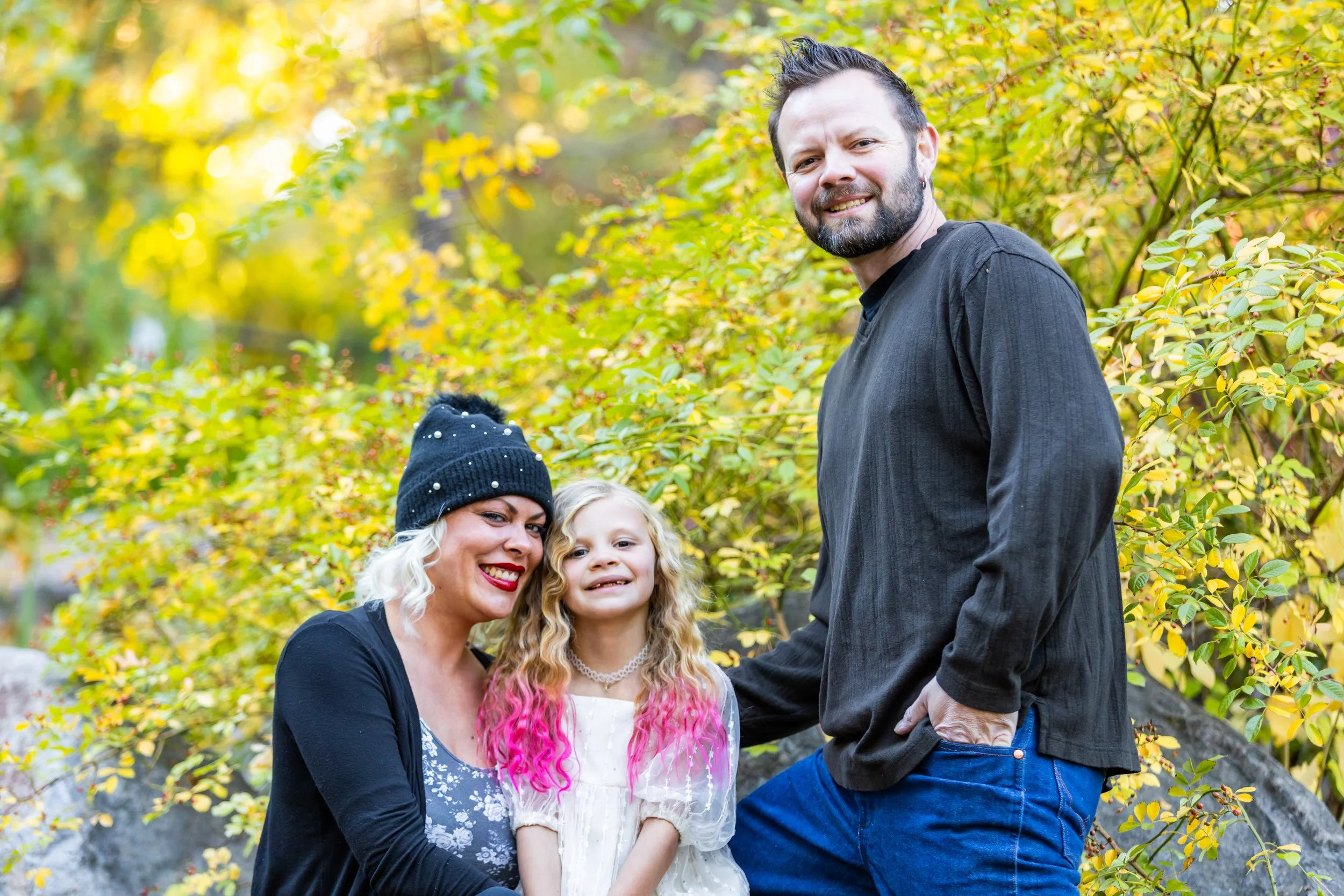 Family portrait with a mother, father, and daughter posing in an autumn setting with lush green foliage. The mother is wearing a black beanie and cardigan, the father is in a dark shirt and jeans, and the daughter is in a white dress with pink-tipped hair.