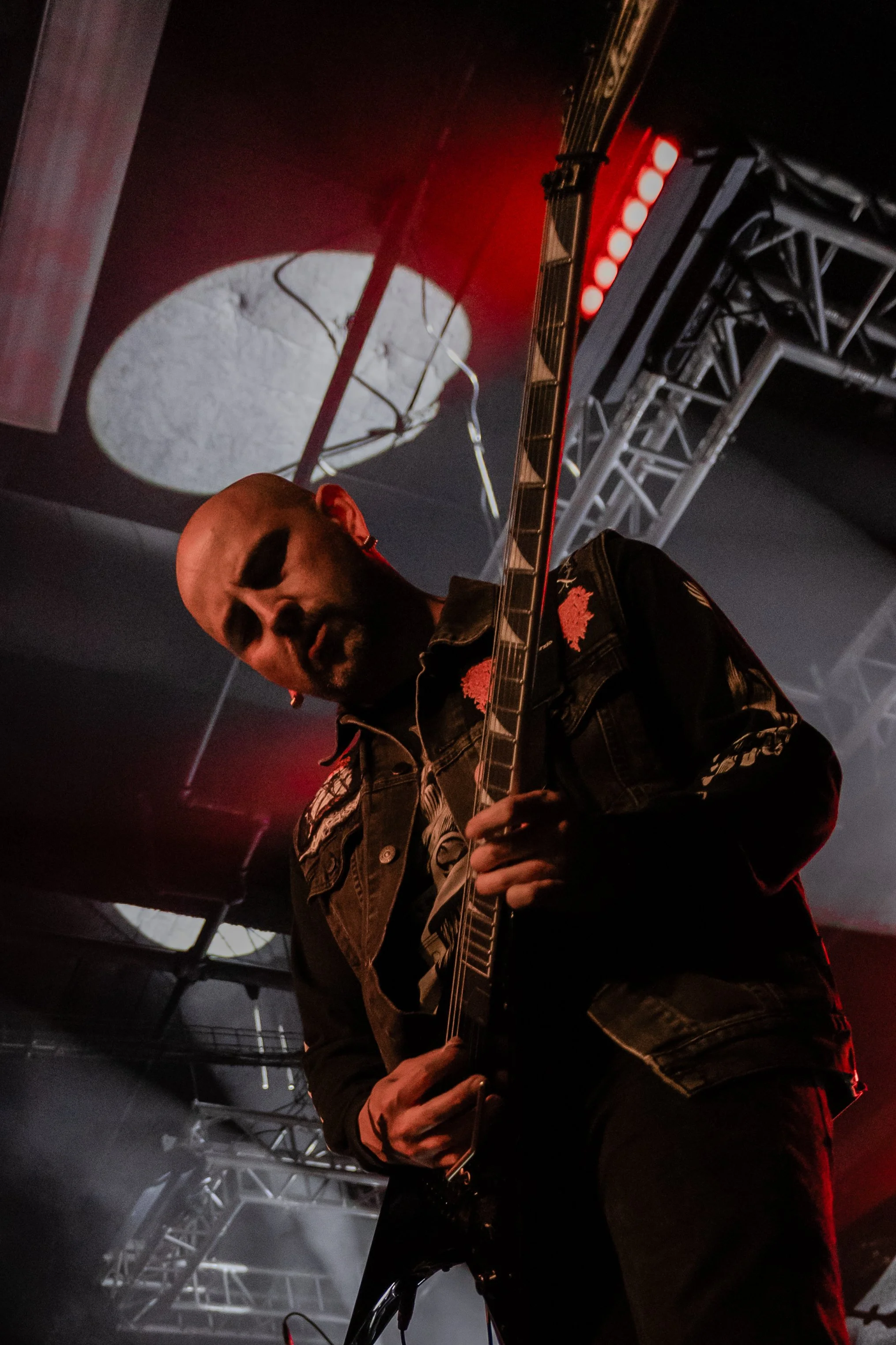 A male guitarist with a shaved head and facial hair playing on stage with a dark background, illuminated by red and white lights, with visible stage equipment and metal trusses overhead.