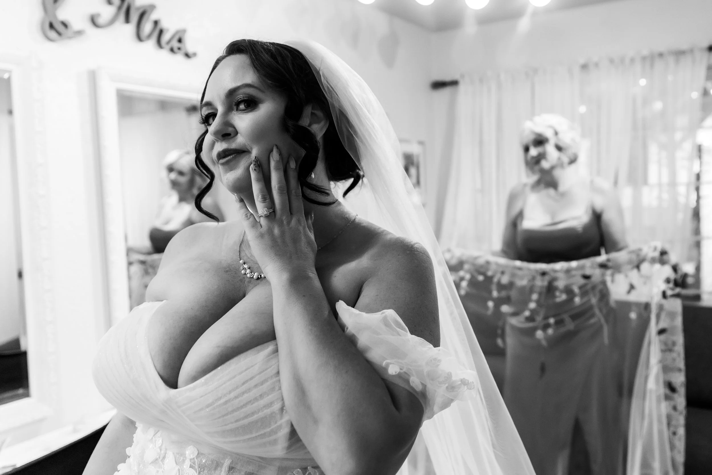 A bride with dark hair in loose curls, wearing a wedding dress and veil, touches her face with her hand, showcasing her engagement ring. Two women are blurred in the background in a room with a mirror and curtains.