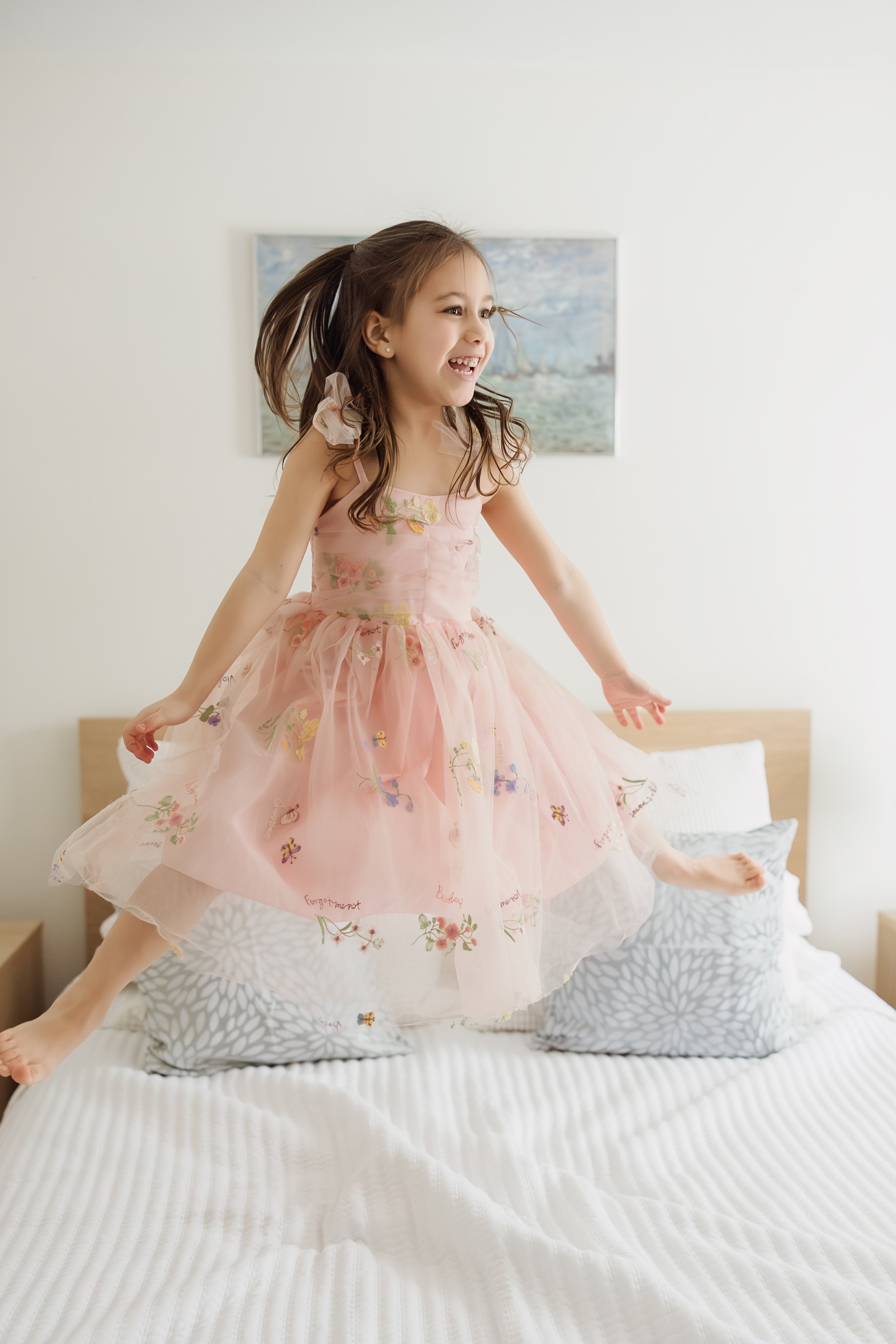 A young girl in a pink floral dress is jumping on a bed with white bedding, in a bedroom with white walls and a painting in the background.