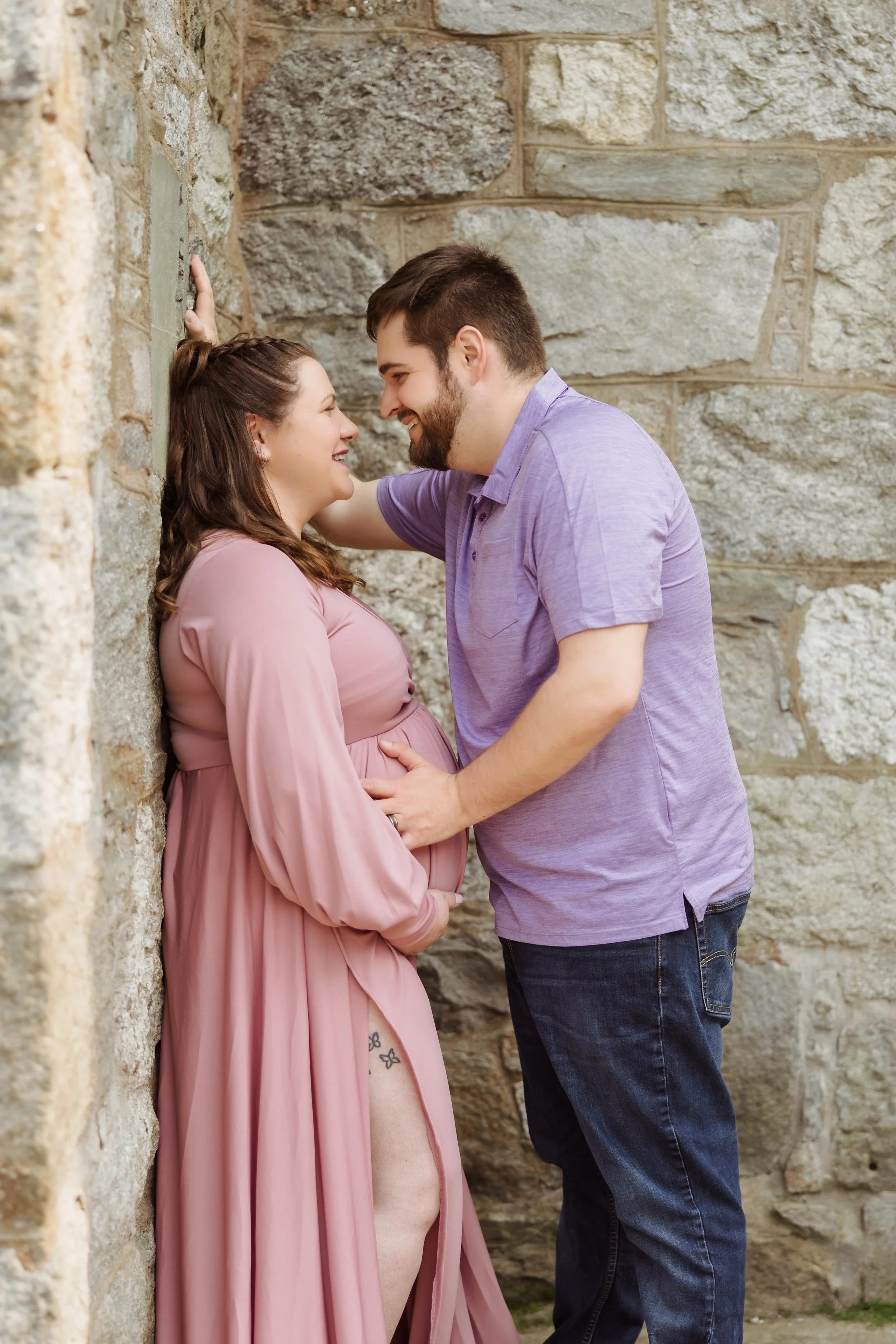 A pregnant woman in a pink dress and the man in a purple shirt are smiling and touching foreheads against a stone wall, sharing an intimate moment.