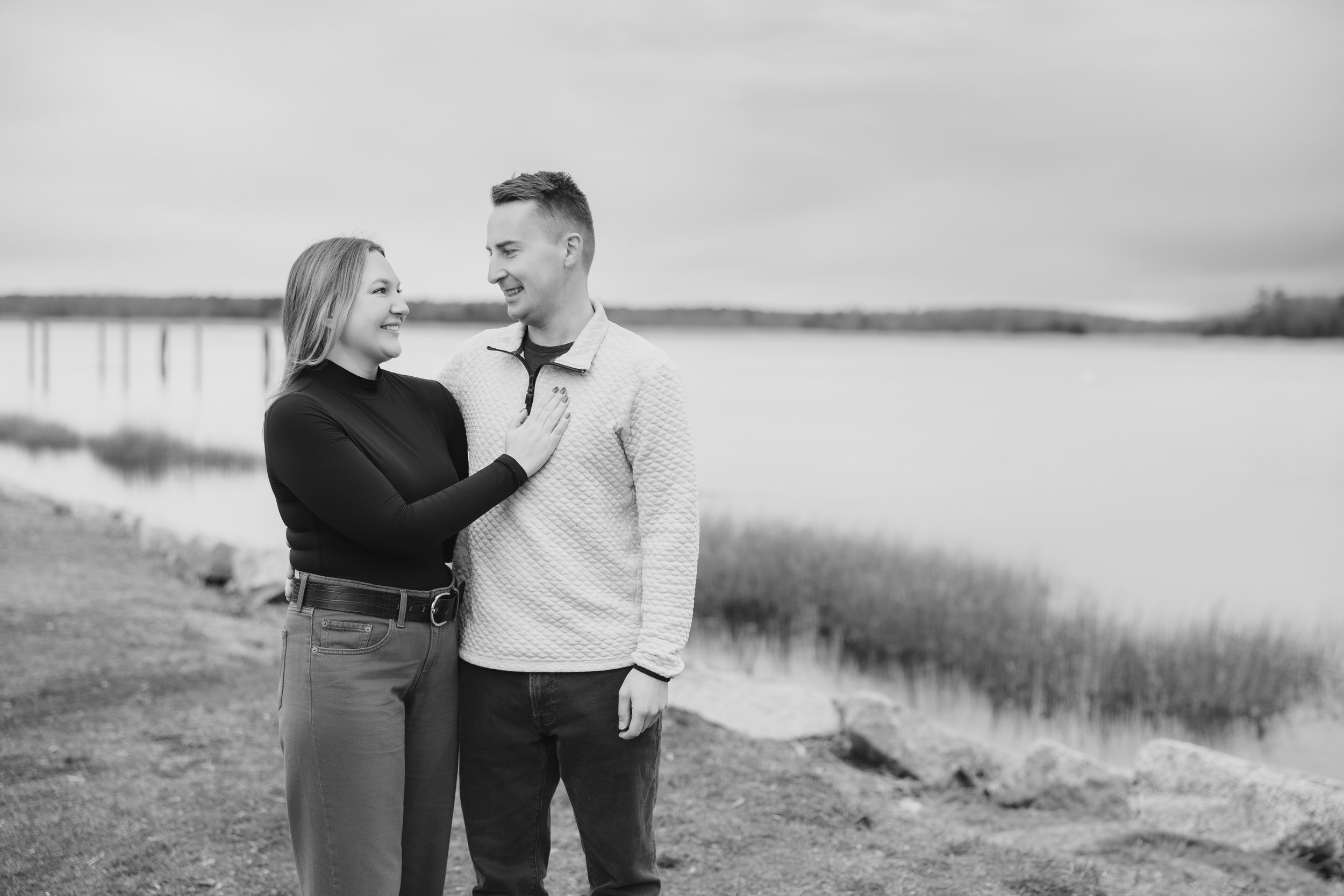 A black-and-white photo of a couple standing near a body of water with a shoreline and cloudy sky in the background, smiling and gazing at each other.