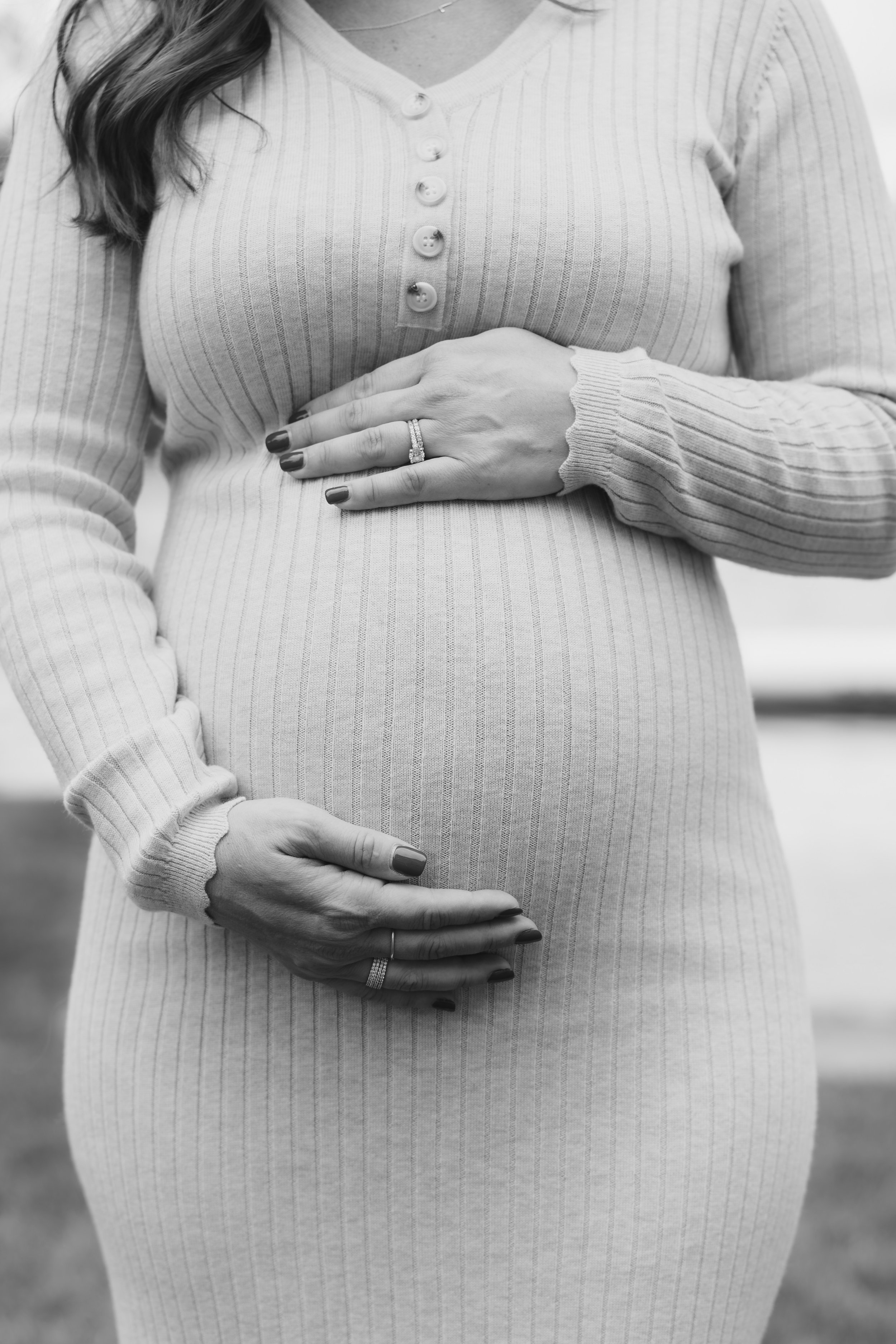 Close-up black and white photo of a pregnant woman wearing a ribbed, long-sleeved shirt with buttons, gently holding her belly with both hands, her left hand showing a wedding ring.