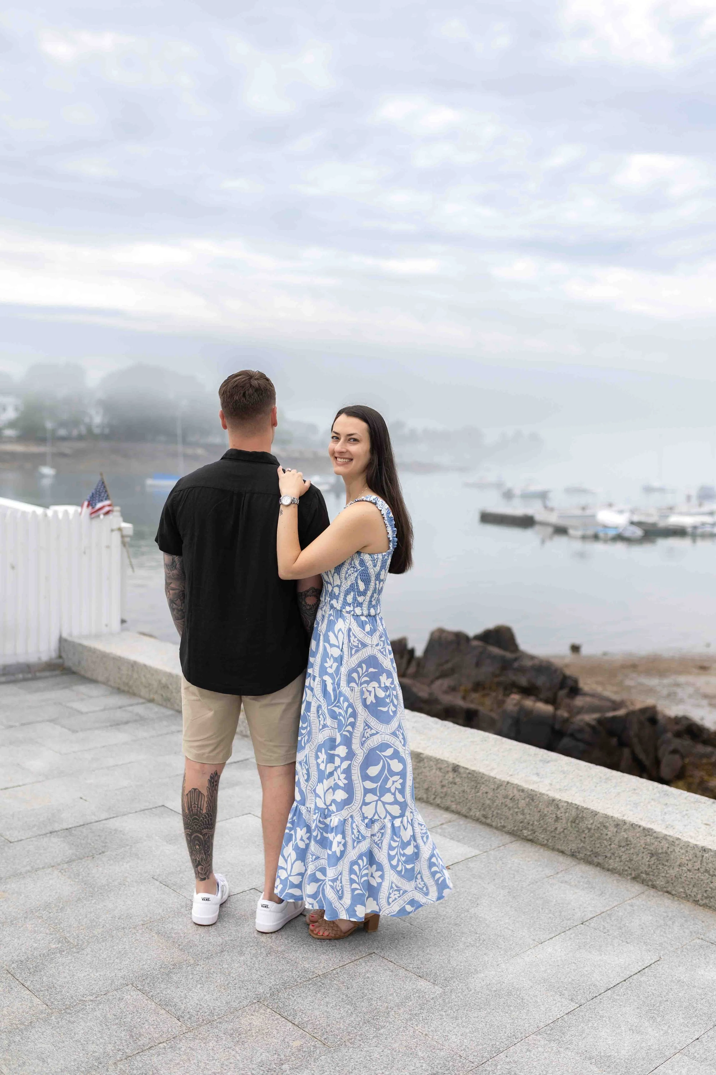 A couple standing on a waterfront promenade, with the woman smiling at the camera. They are near a body of water with boats and a foggy sky in the background. The woman is wearing a long blue and white patterned dress, and the man is dressed casually