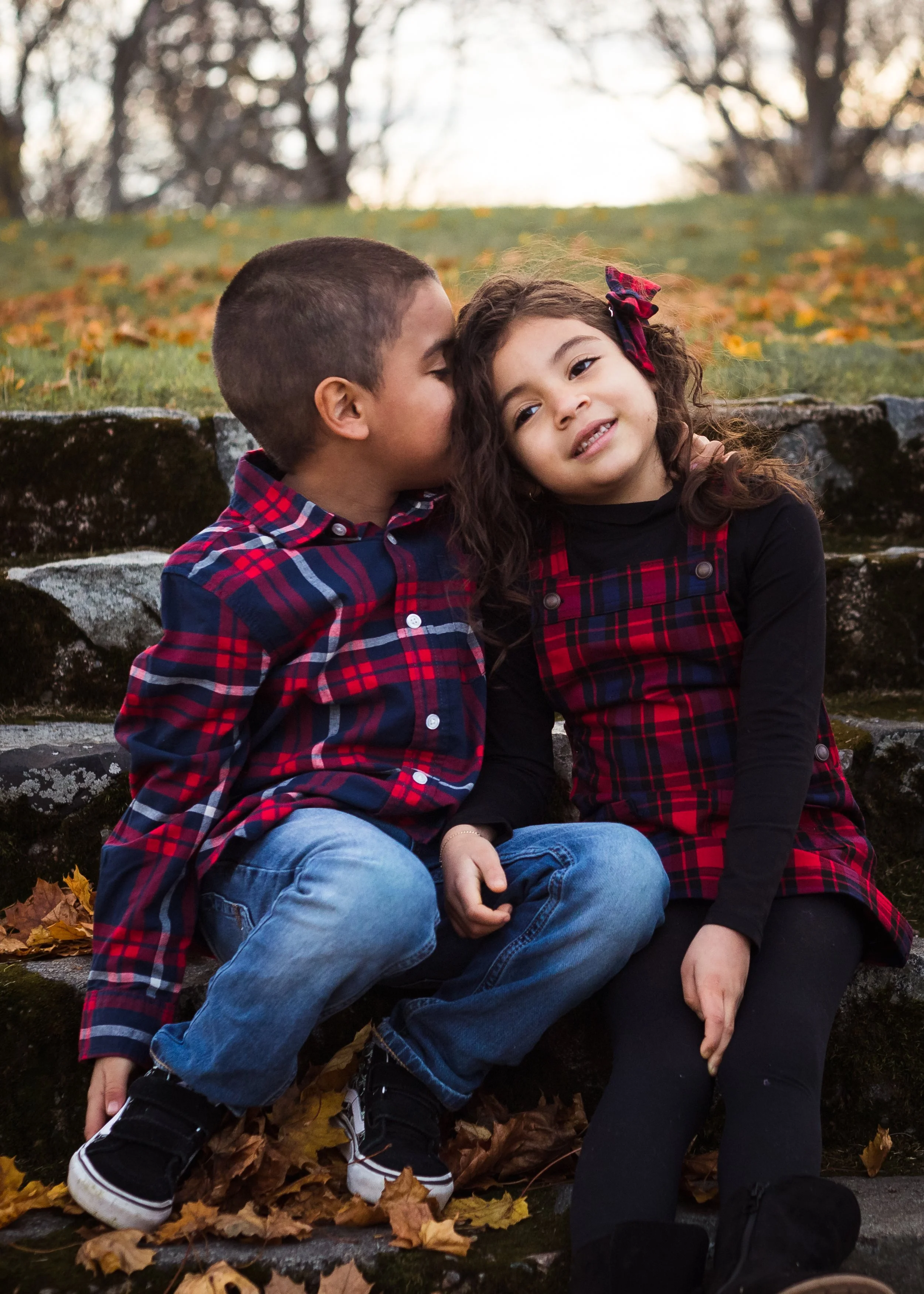 Two children, a boy and a girl, sitting outdoors on a stone ledge with autumn leaves on the ground. The boy is kissing the girl's cheek, and they are both smiling. They are wearing plaid shirts, and the girl has a bow in her hair.