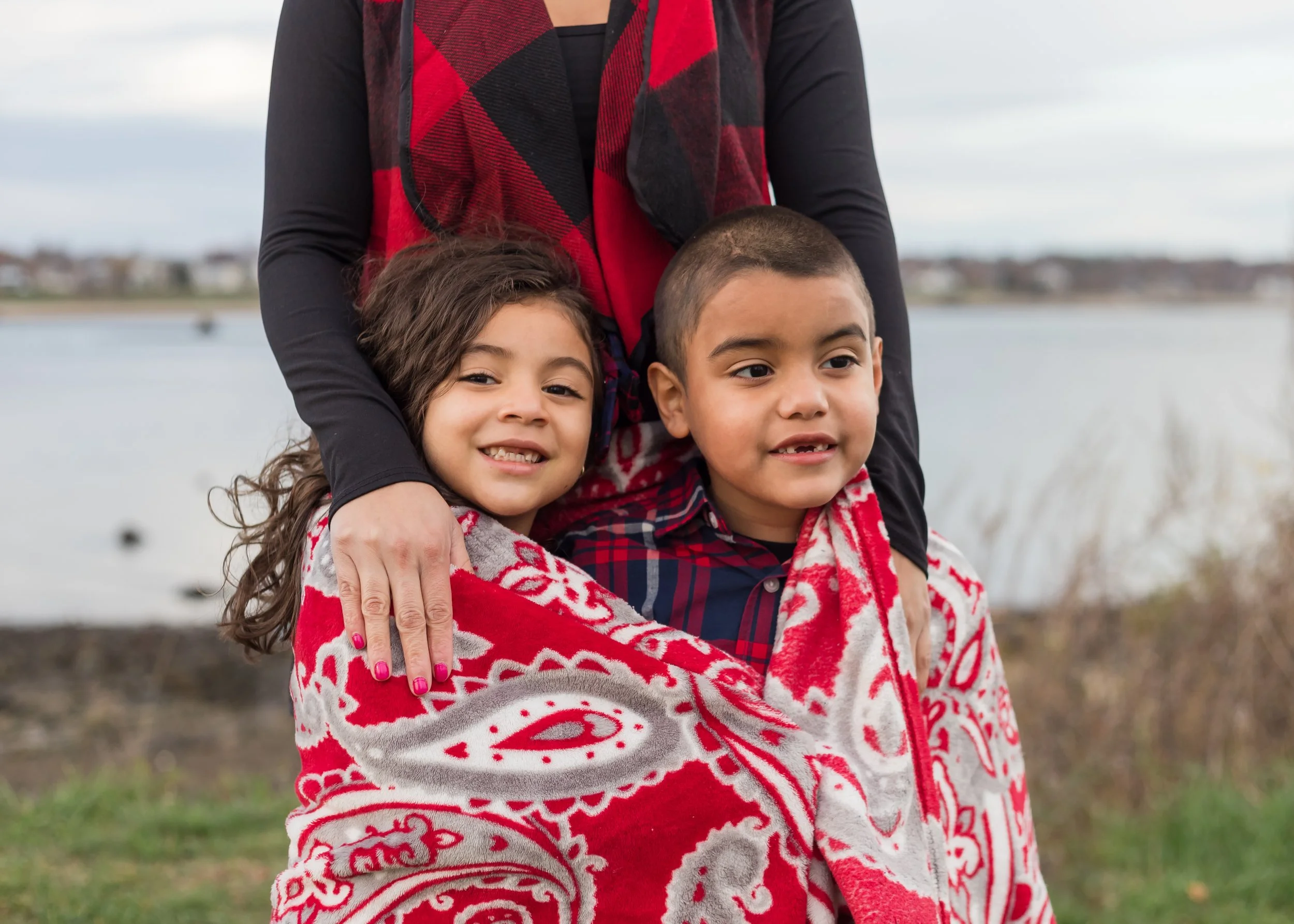 A woman with two children outdoors near a body of water, wearing a black shirt and a red and black plaid scarf. The children are wrapped in red and white patterned blankets.
