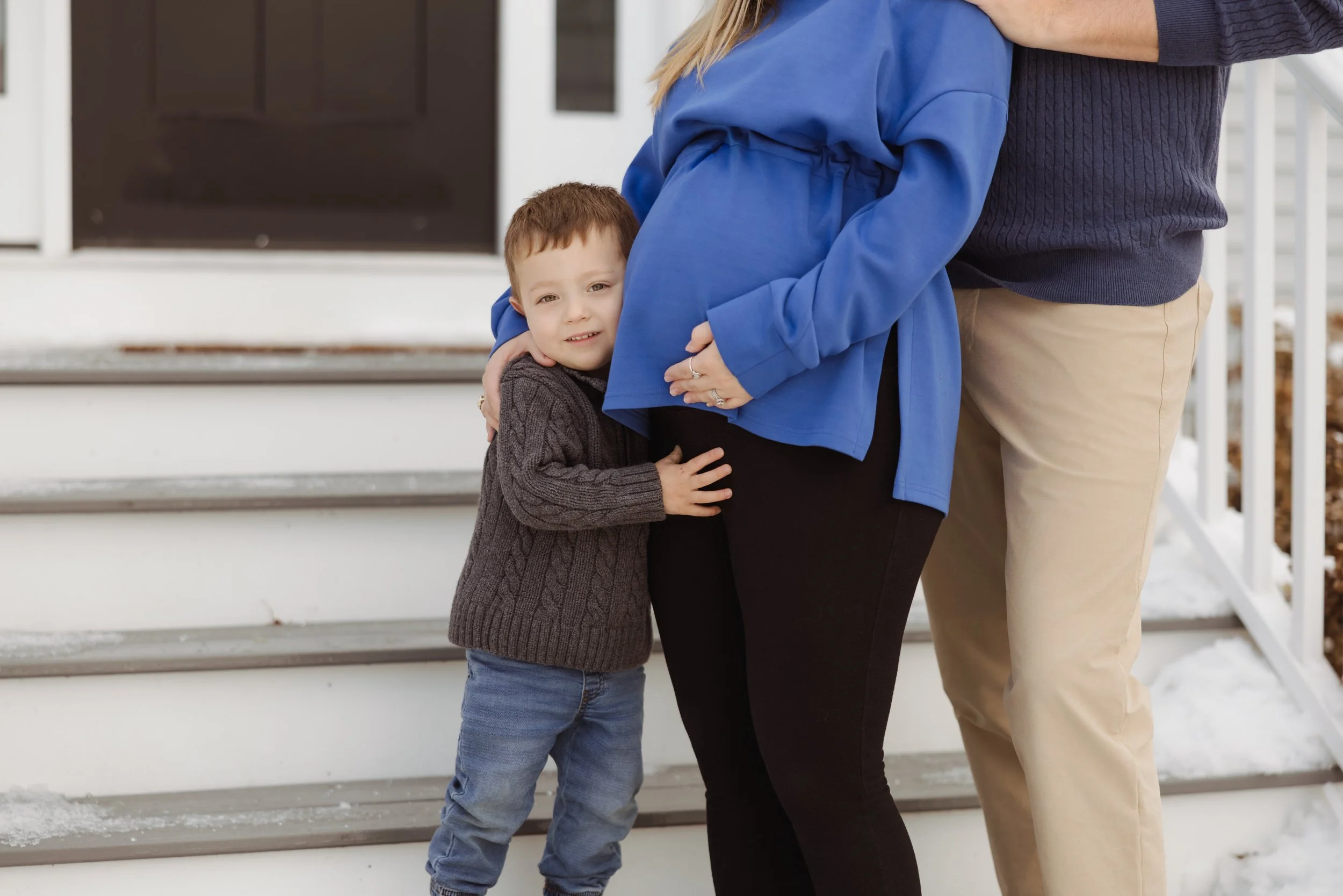 A young boy hugging a woman outside on the front porch of a house, with another person standing nearby. The boy has short brown hair, and the woman is wearing a blue jacket. The setting is winter with snow on the stairs.