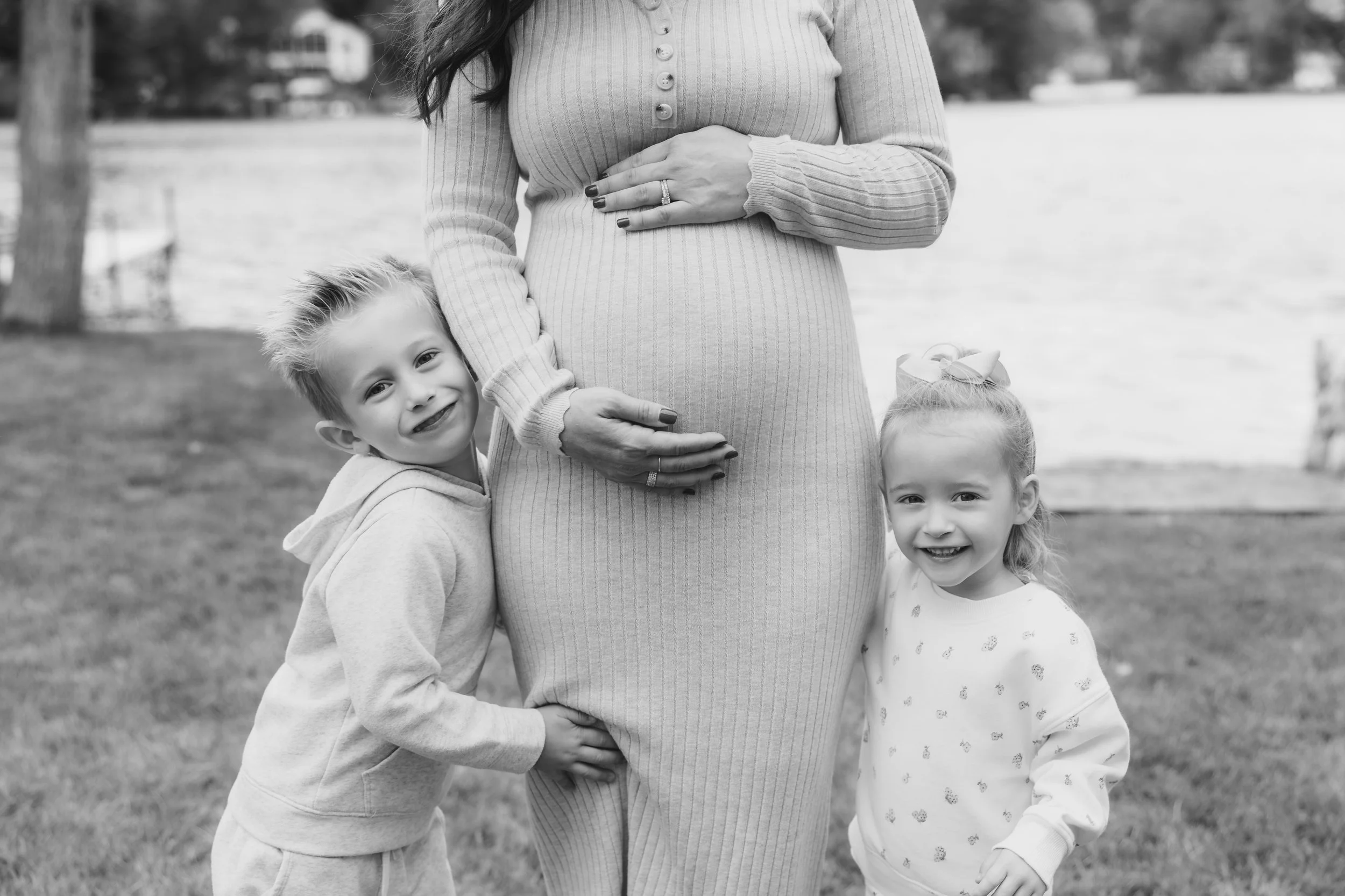 A pregnant woman standing outdoors near a lake with her two young children, a boy and a girl, hugging her belly. The woman is wearing a long-sleeve, buttoned sweater, and the kids are smiling, with the girl smiling at the camera. The image is in blac