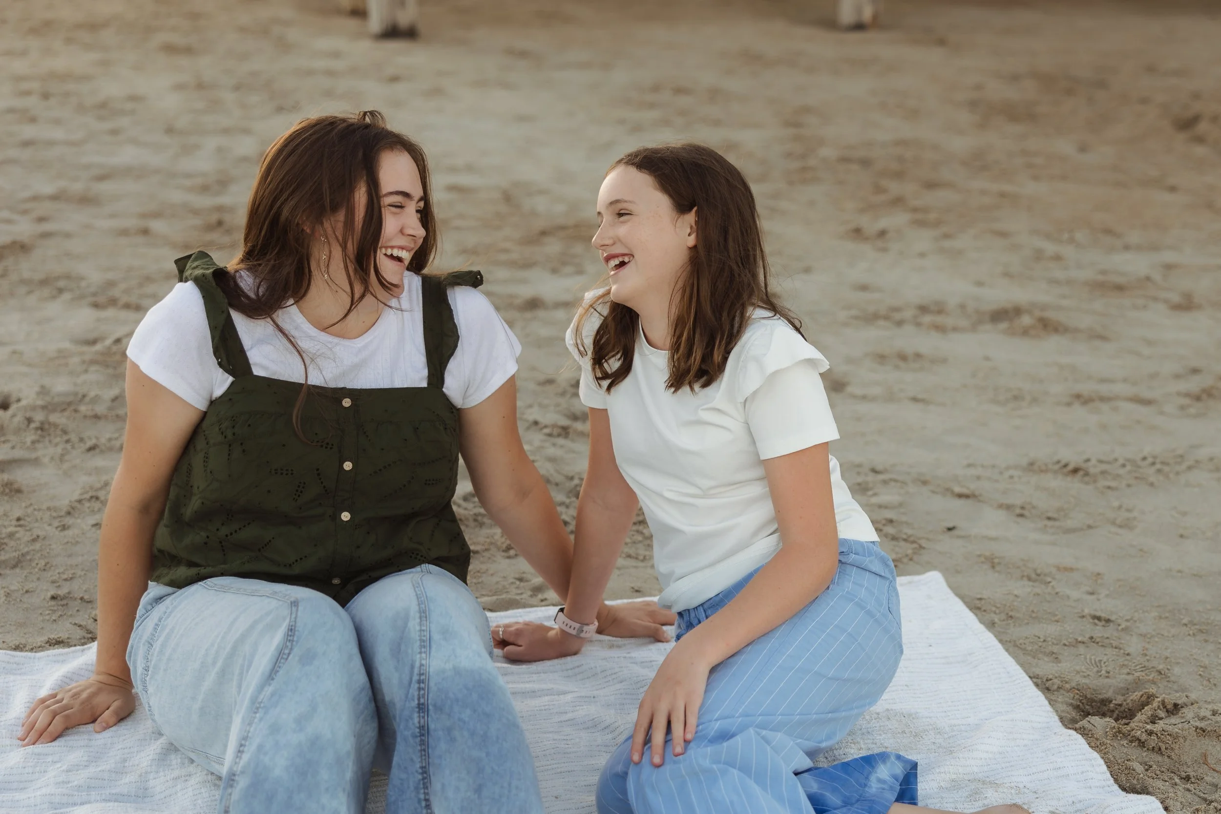 Two girls sitting on a beach towel, smiling and laughing together.