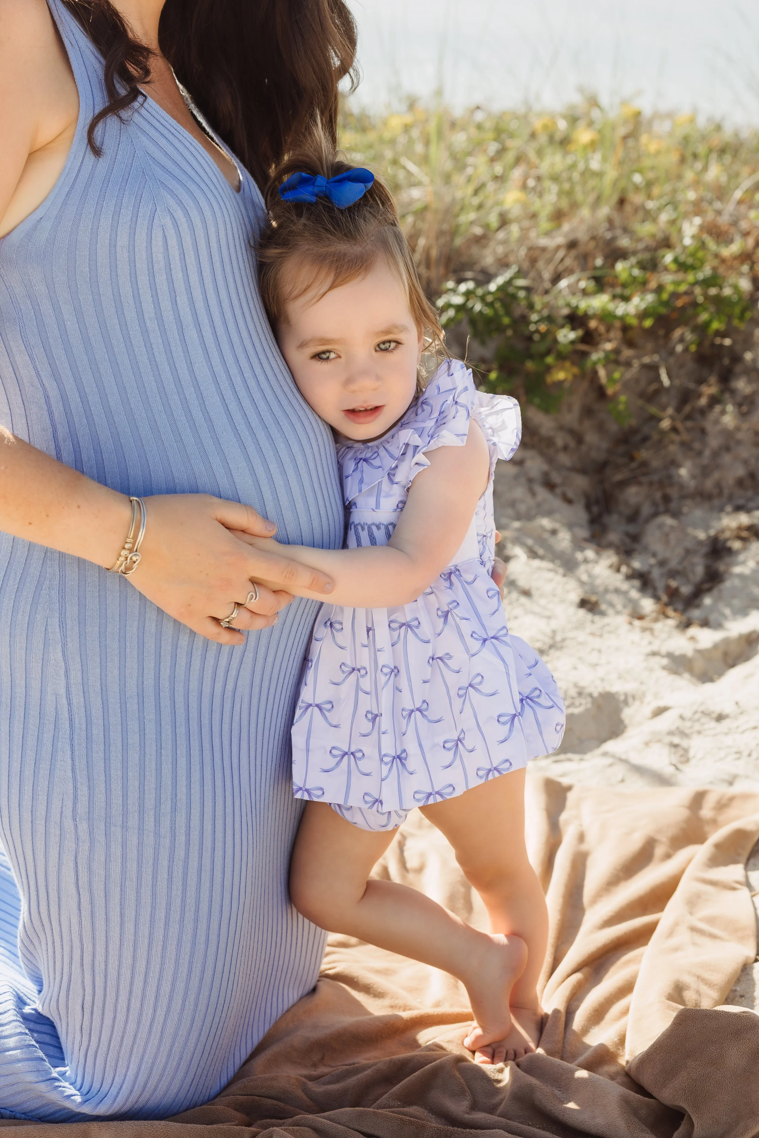 A woman and a young girl standing on a sandy beach. The woman is wearing a blue dress, and the girl is wearing a white dress with purple bows and a blue bow in her hair. The woman is holding the girl, who is standing on her leg. In the background, th