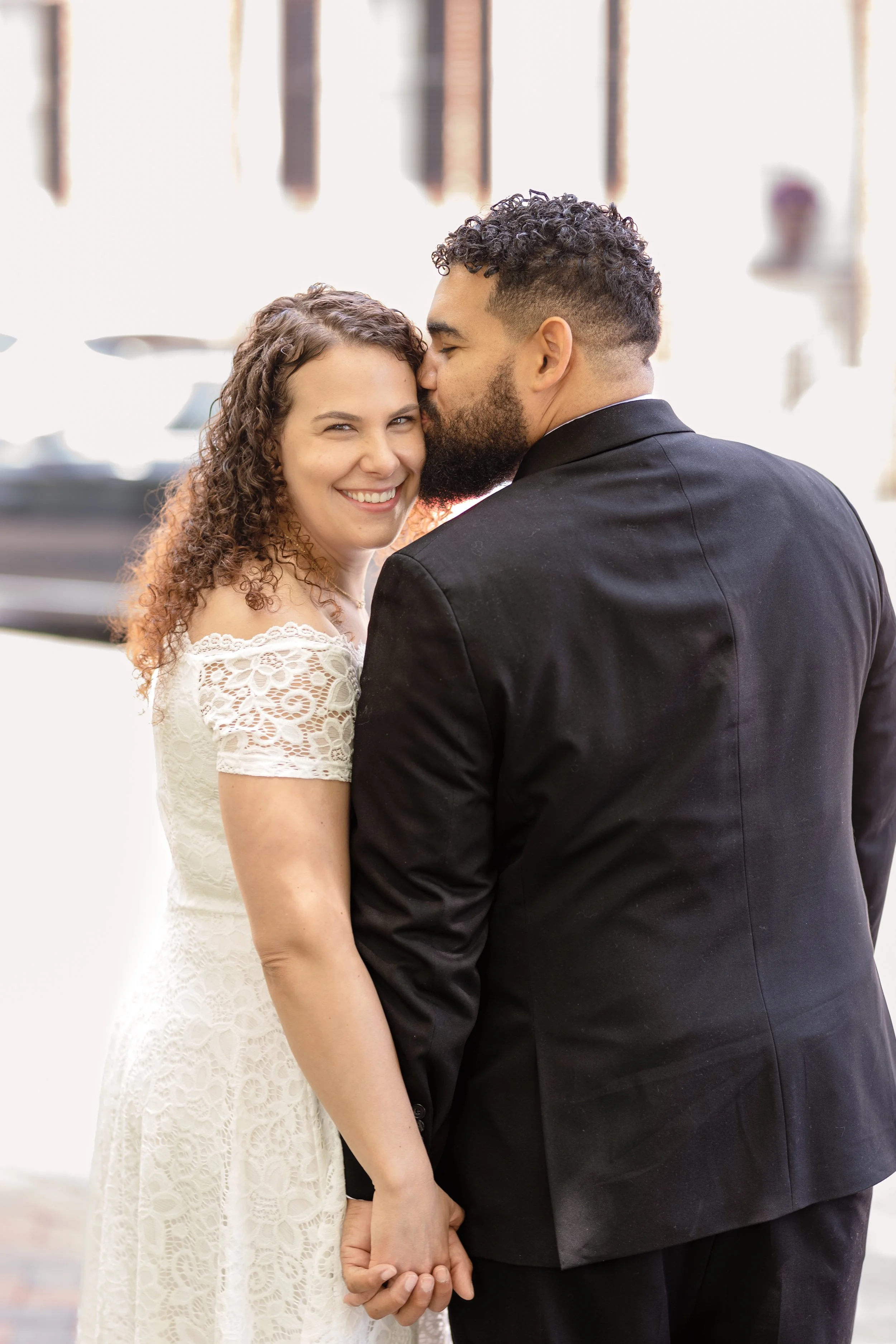 A couple, a woman with curly hair wearing a white lace dress and a man with short curly hair and a beard in a black suit, share a kiss on the forehead while holding hands, smiling. The background is bright and slightly blurred.