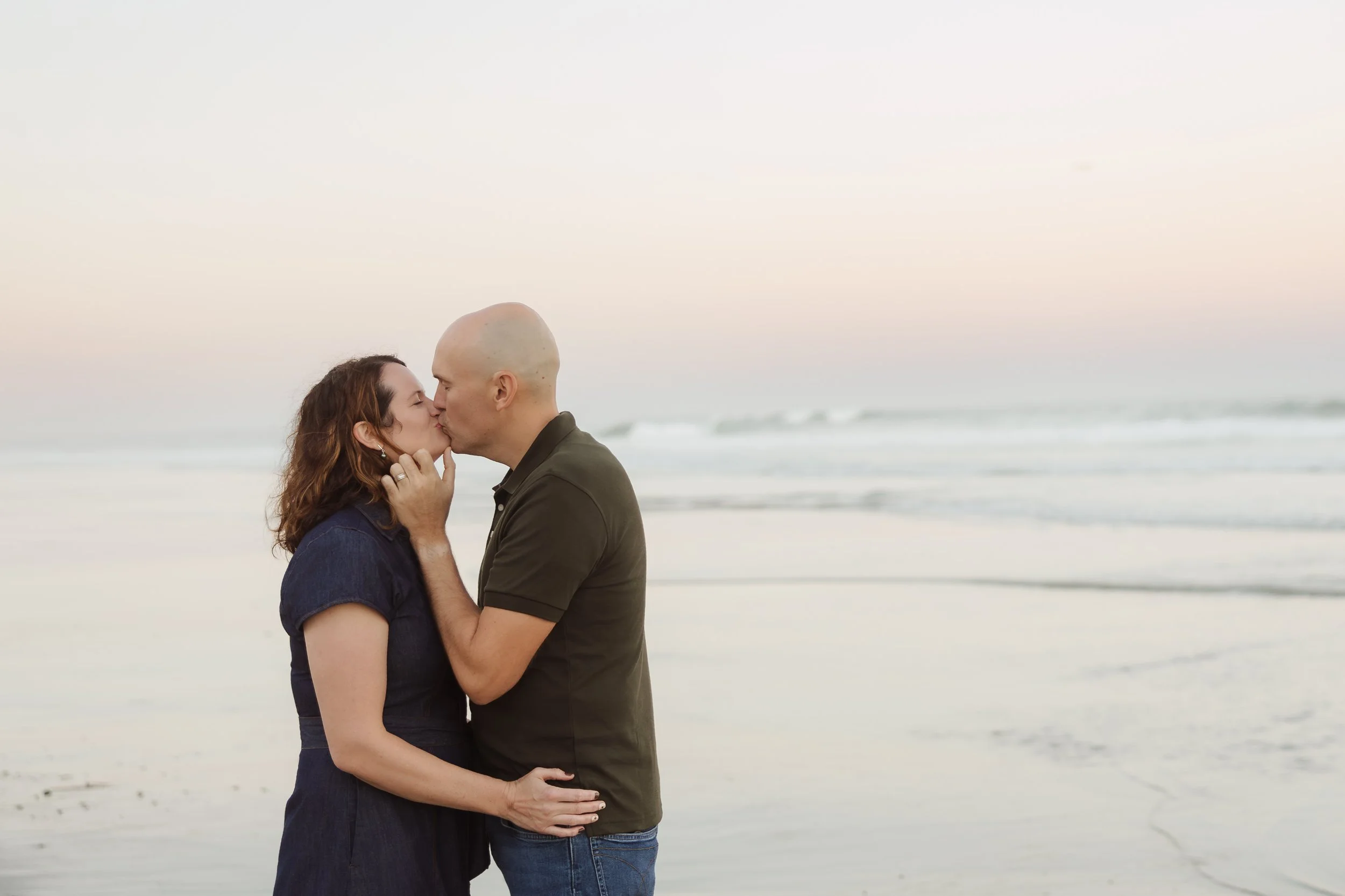 A couple sharing a kiss on the beach during sunset, with the ocean in the background.