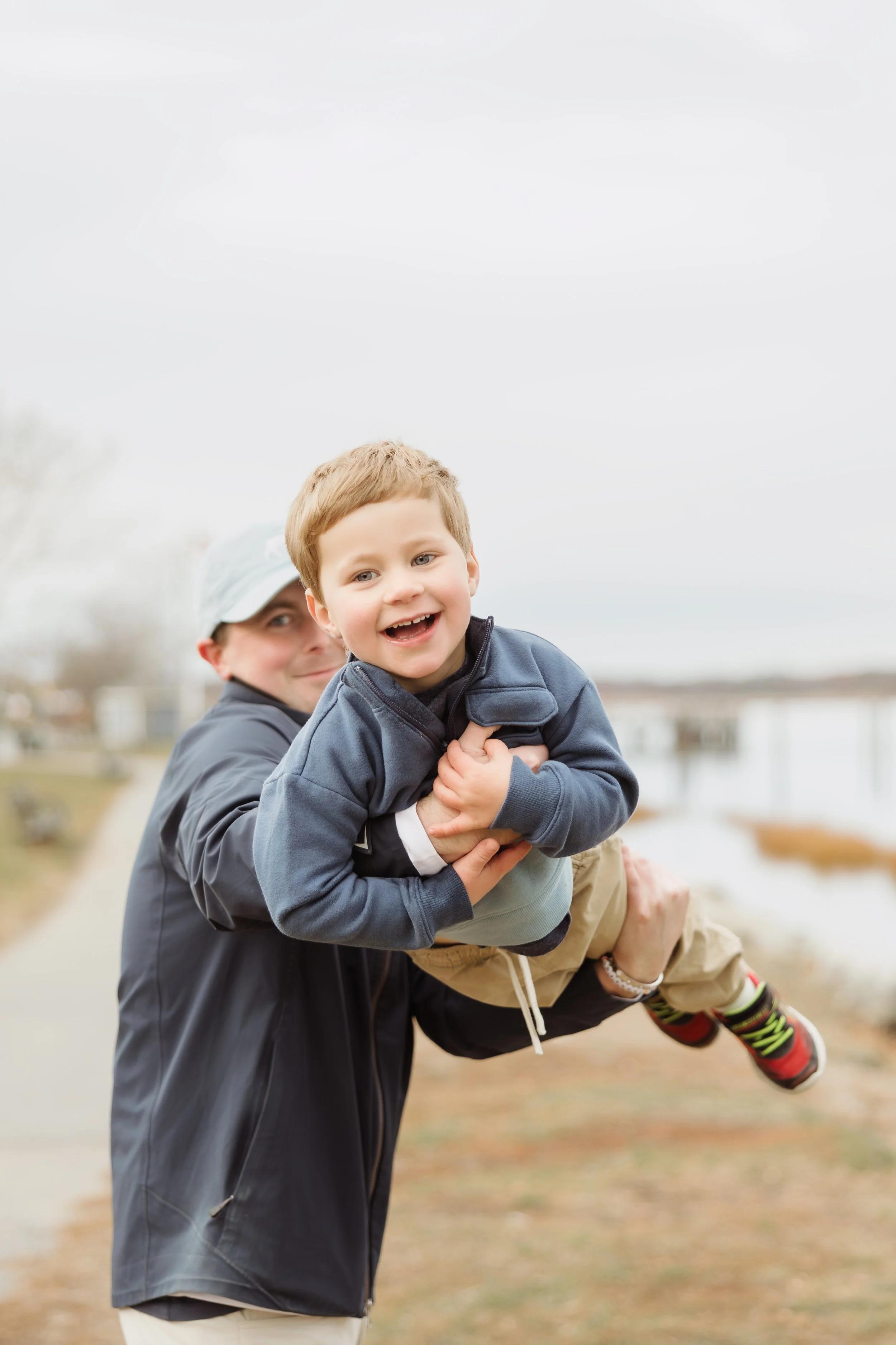 A young boy with curly hair smiling and being held up by an adult man outdoors near a body of water. The man is partially visible, wearing a cap and jacket, and the background is slightly blurred with overcast skies.