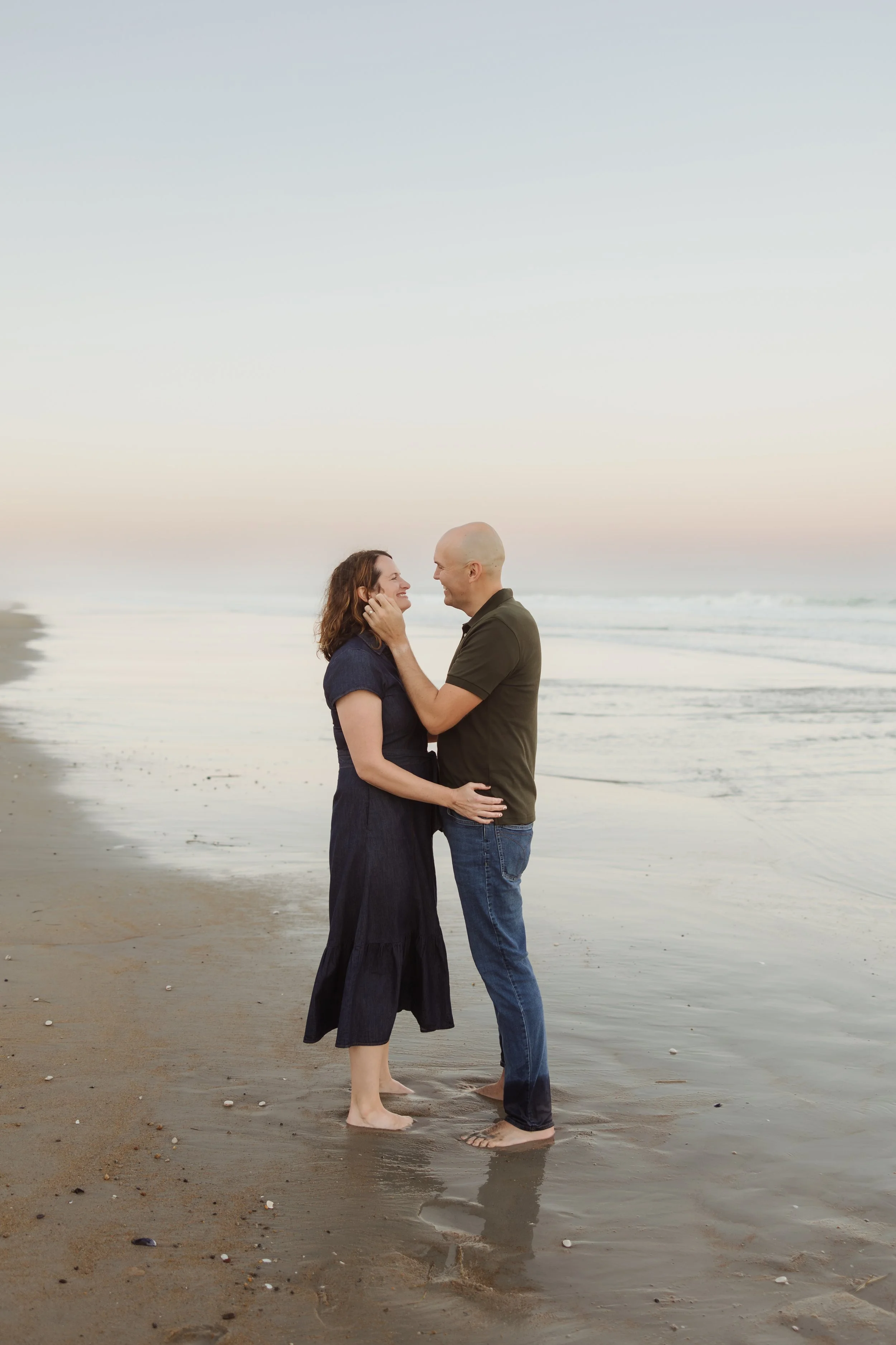 A couple standing on a beach at sunset, facing each other and smiling, with the ocean in the background.