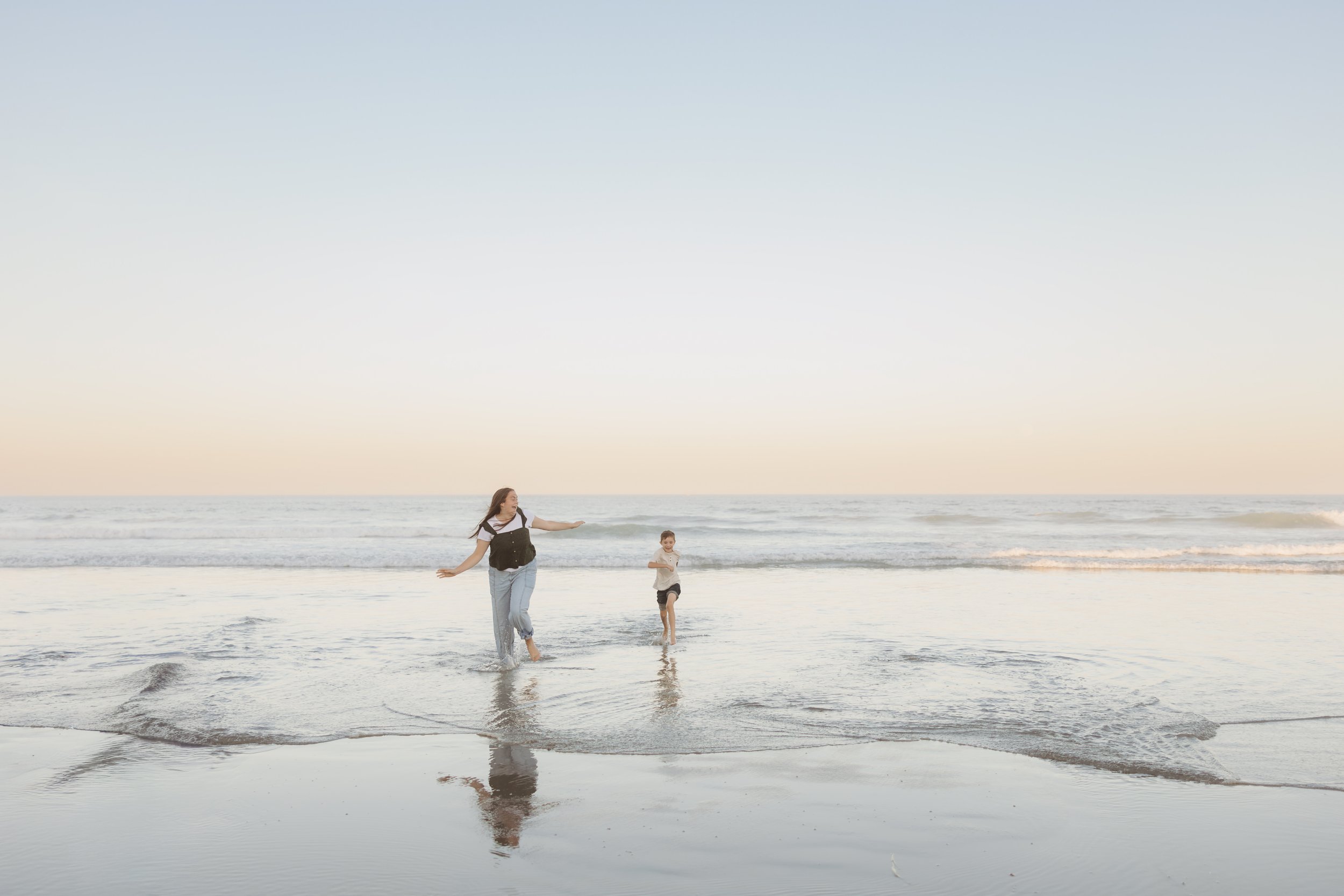 A woman and a young child run through shallow ocean water at sunset, with the sea and sky in the background.