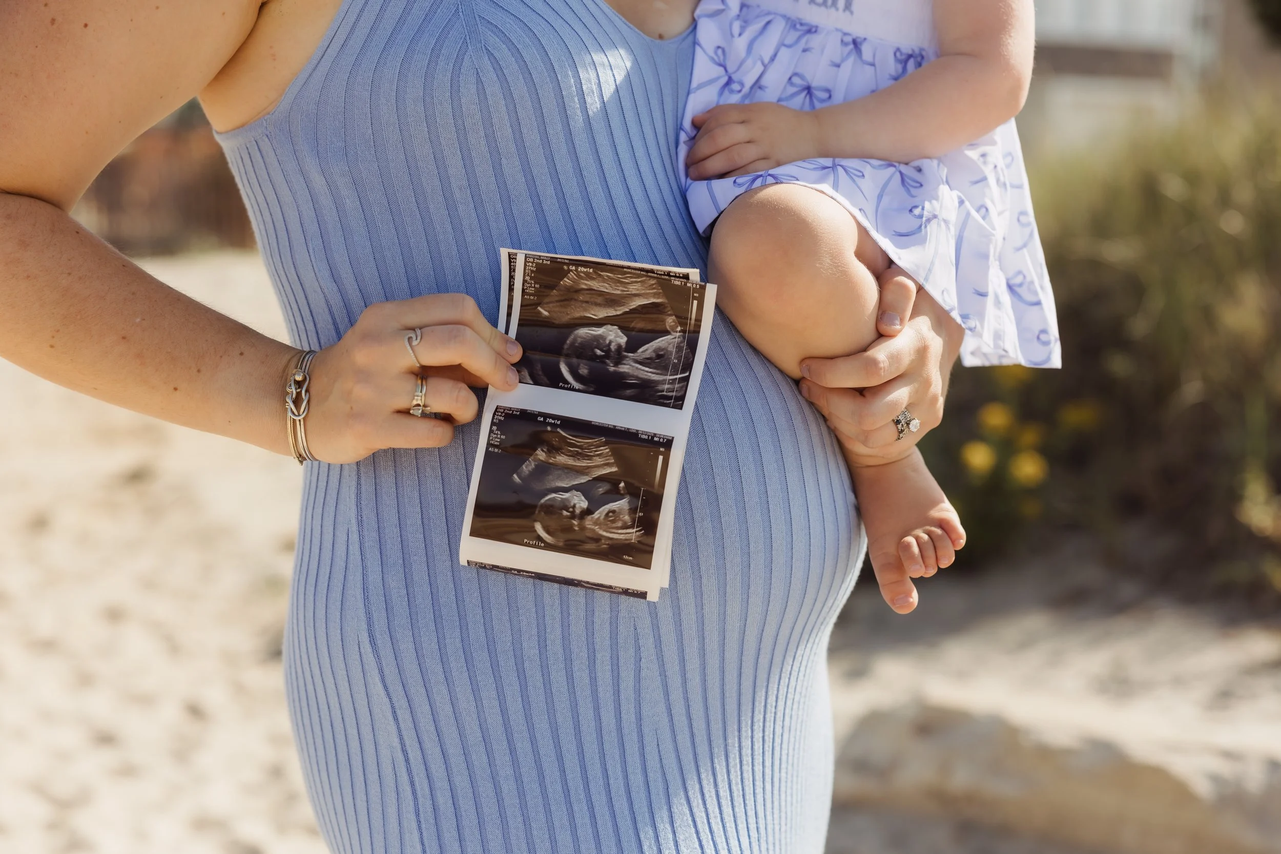 A pregnant woman holding ultrasound pictures on her belly, standing outdoors on a sunny day.