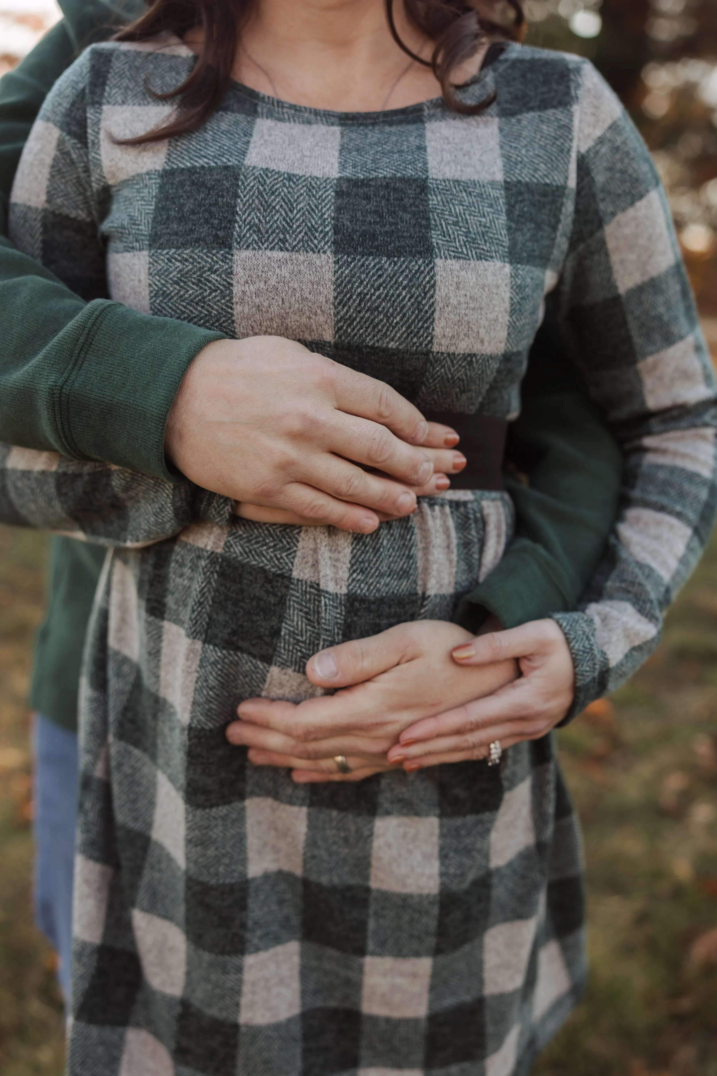 Two women are hugging outdoors, one standing behind the other with her hands placed on the woman's stomach, suggesting a pregnancy. The woman in front is wearing a plaid shirt with shades of gray, black, and tan, while the other is wearing a green lo