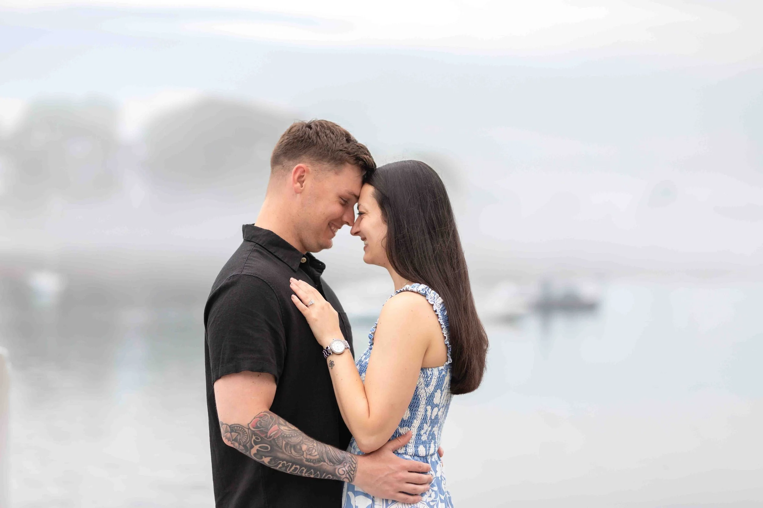 A couple stands close, smiling and touching foreheads. They are outdoors with a cloudy sky in the background.