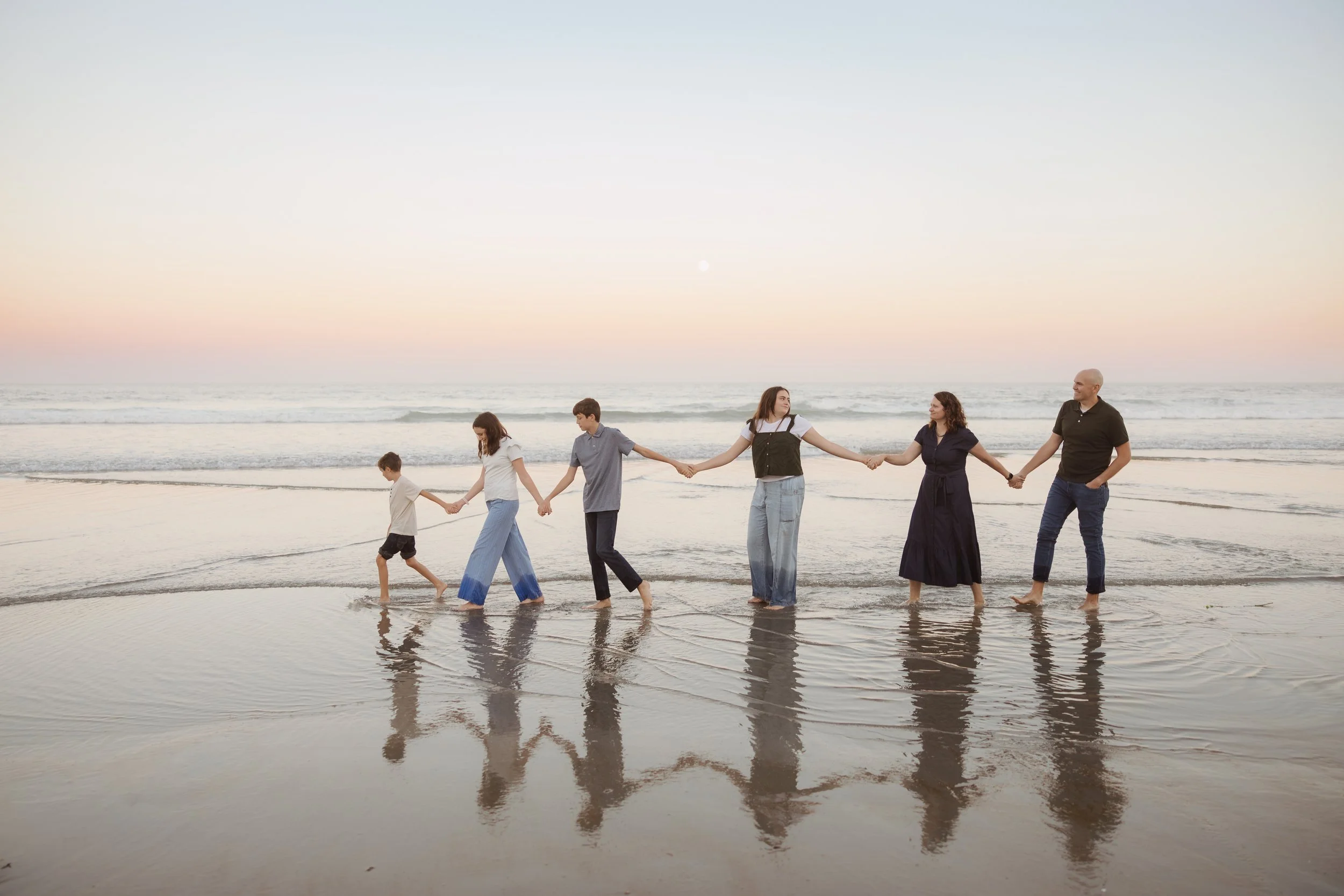 Family of six walking hand in hand along the beach during sunset, with reflections visible in the shallow water.