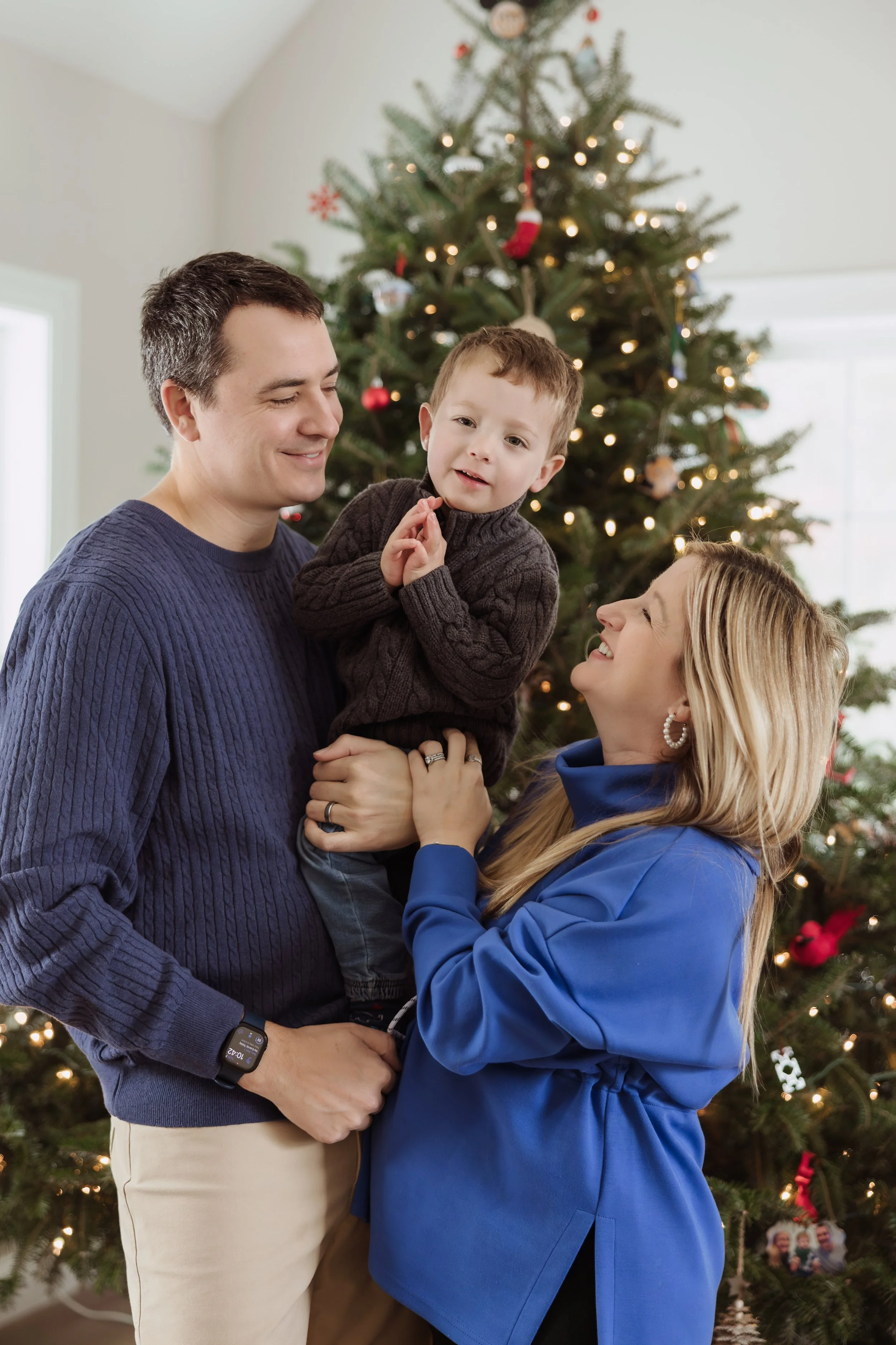 A family of three celebrating Christmas in front of a decorated tree. The father, wearing a navy sweater, is holding a young boy in a brown sweater. The mother, in a blue jacket, is smiling at them.