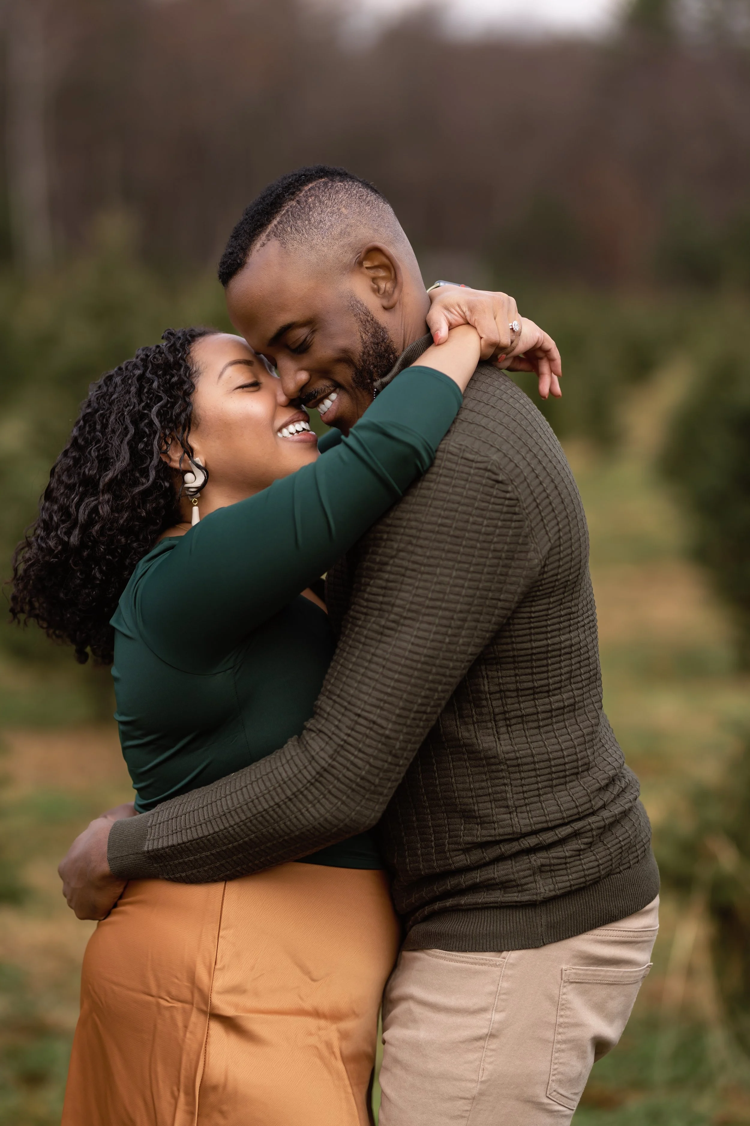 A couple embracing outdoors during autumn, smiling with their noses touching, the woman with curly hair and earrings, and the man with a short haircut and beard.