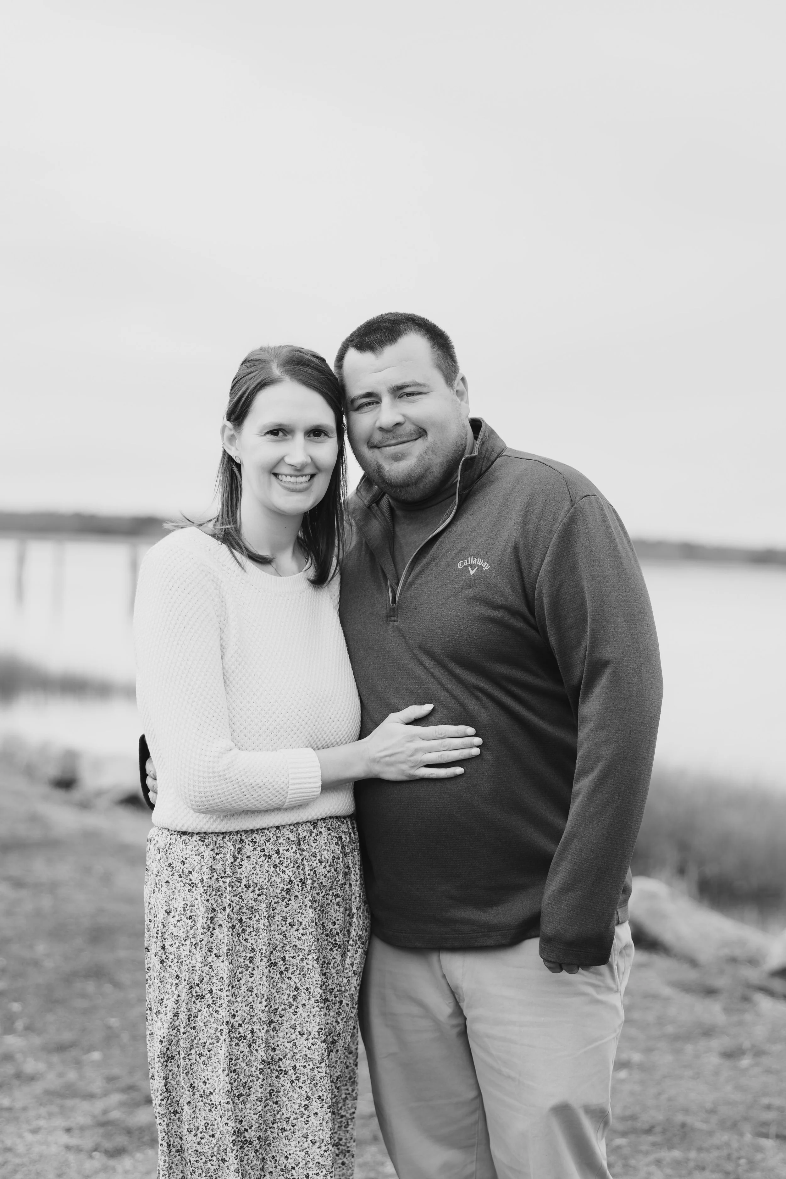 A smiling woman and man standing outdoors by a body of water, embracing each other with the woman's arm around the man's waist and her hand resting on his stomach.
