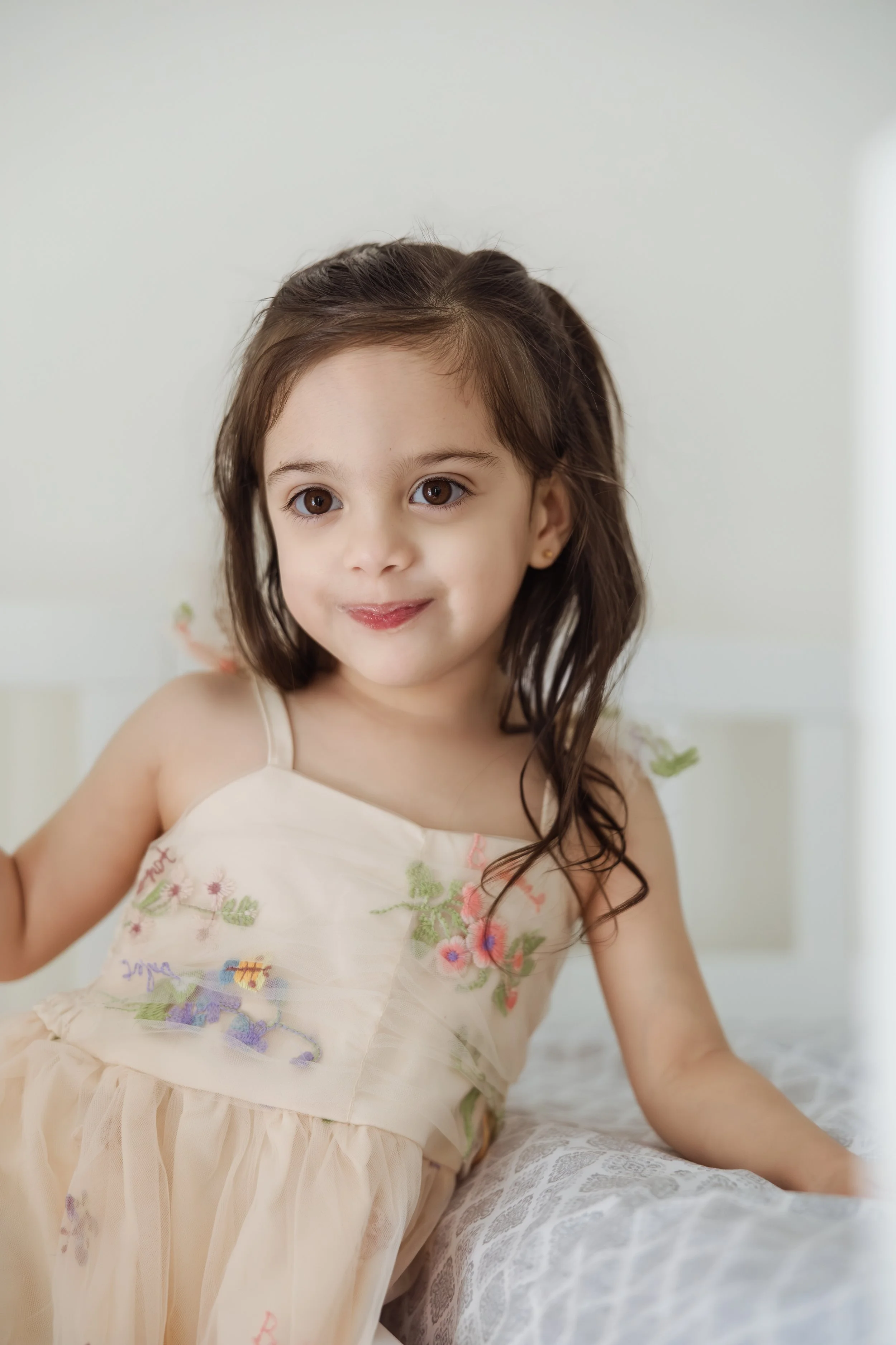 A young girl with brown hair and brown eyes smiling softly, wearing a light-colored dress with floral embroidery, sitting on a bed with a patterned sheet, in a softly lit room.