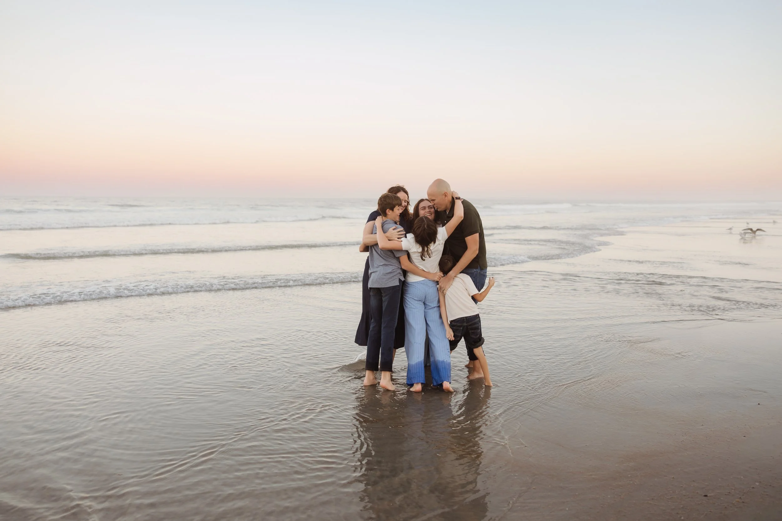 A family of six enjoying time together at the beach during sunset, standing in the shallow water and hugging each other.