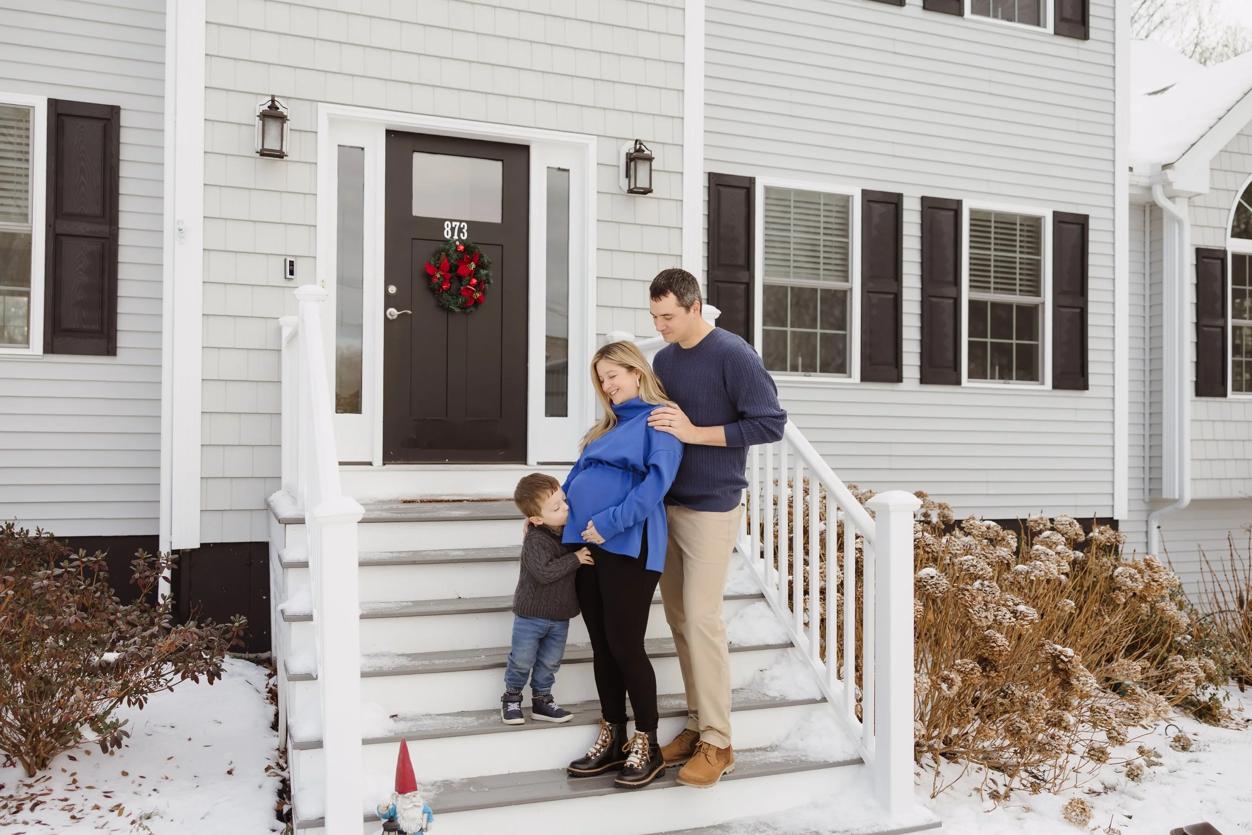 A family of three standing on the front steps of their house during winter. The house has a dark front door decorated with a holiday wreath, white siding, and multiple windows with dark shutters. Snow is on the ground and on the bushes, and the girl 
