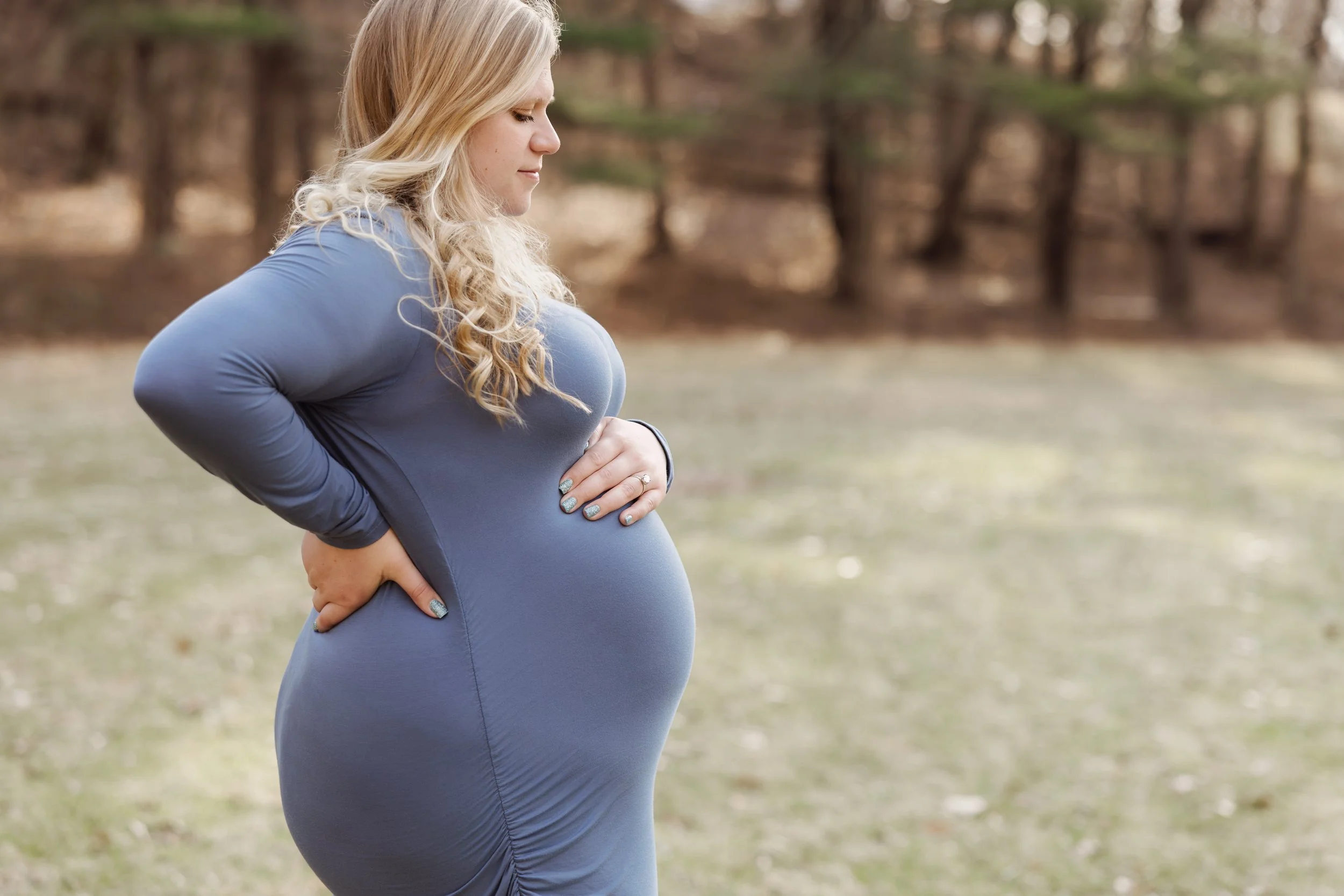 Pregnant woman standing outdoors with her hands on her belly, in a profile view, wearing a blue long-sleeve top, with blurred trees in the background.