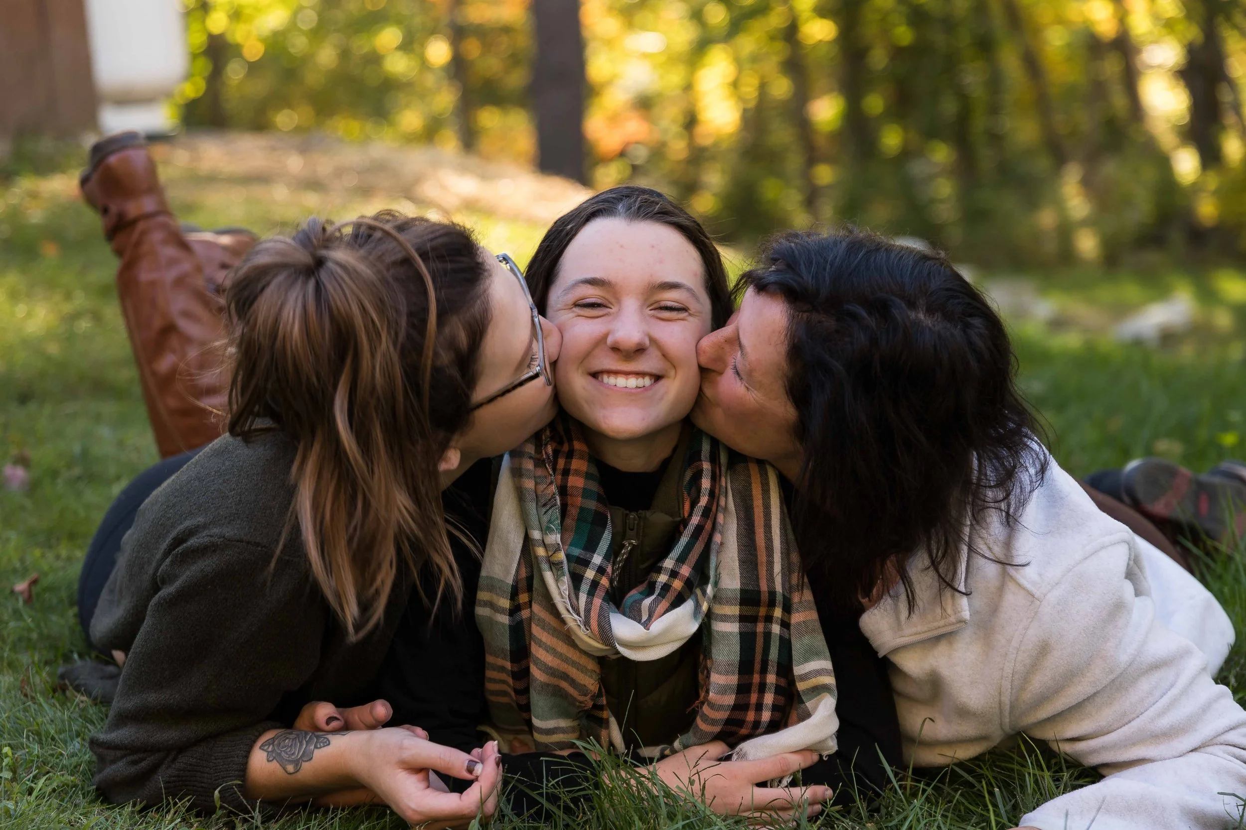 Three smiling young women lying outdoors on the grass, with two women kissing the girl in the middle on the cheeks. The background has trees and a sunny sky.