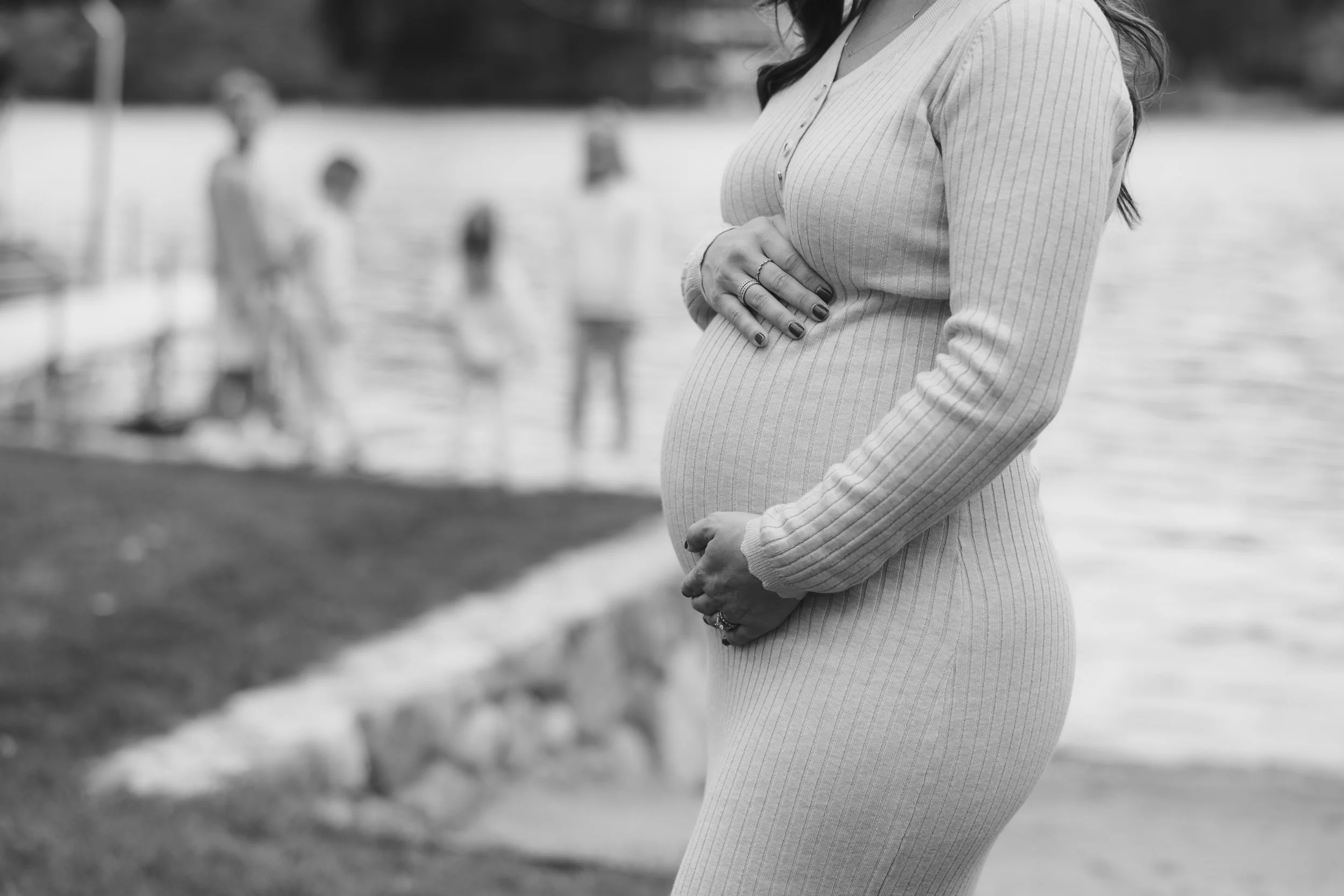 Black and white photo of a pregnant woman standing outdoors near water, with her hands gently resting on her belly, wearing a long-sleeve ribbed top. Blurred figures of people are visible in the background.